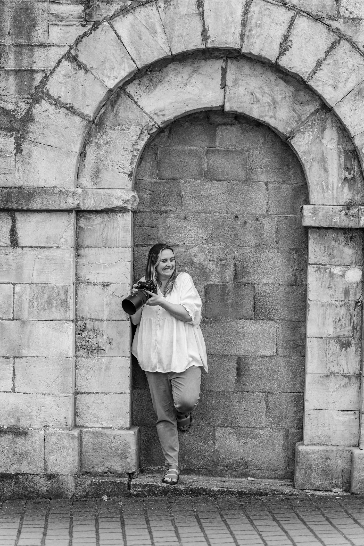 Woman with camera smiling in stone archway, black and white photography portrait.