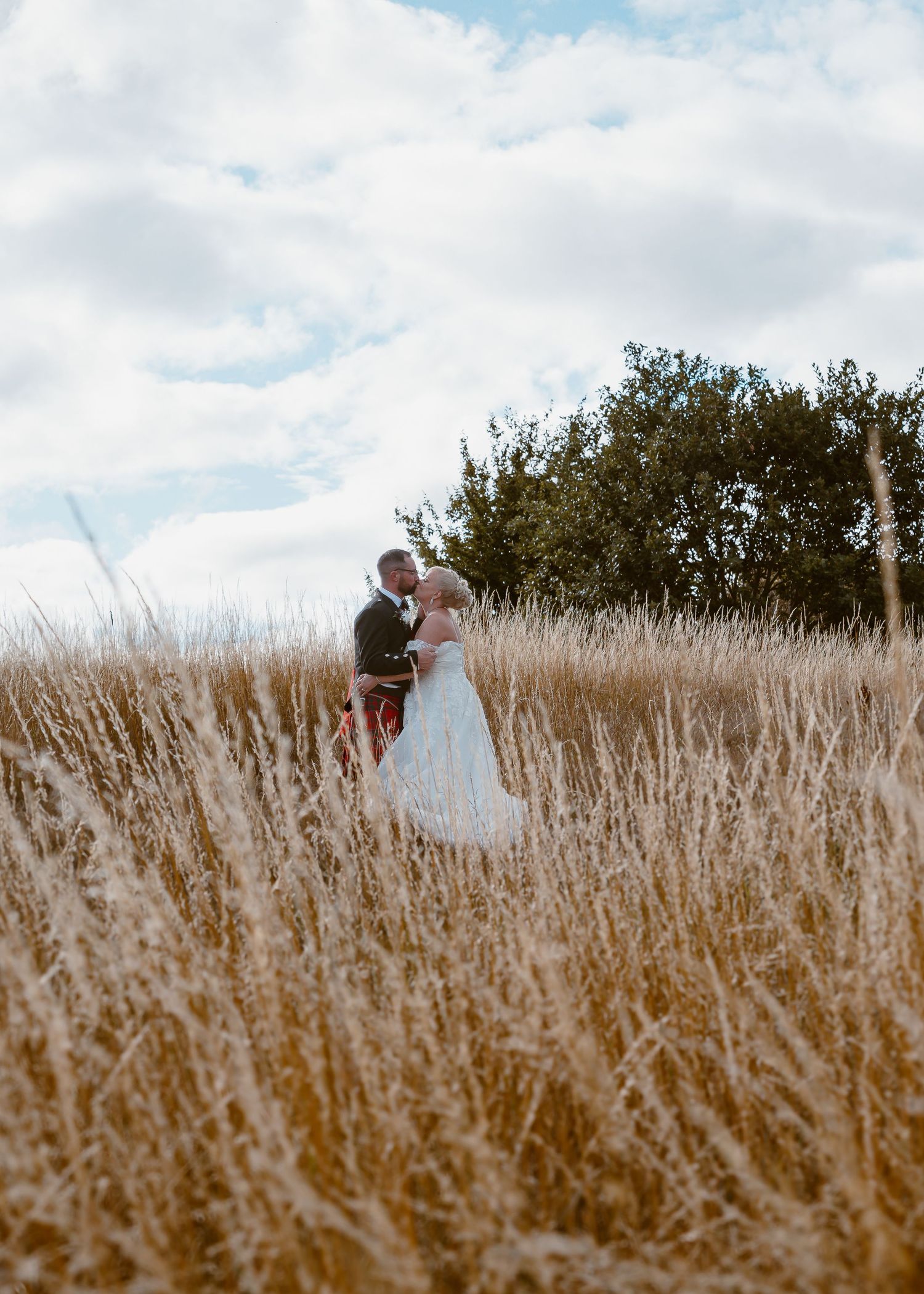 Waterton Park Hotel Wedding A couple stands together in a field of tall golden wheat against a cloudy blue sky.