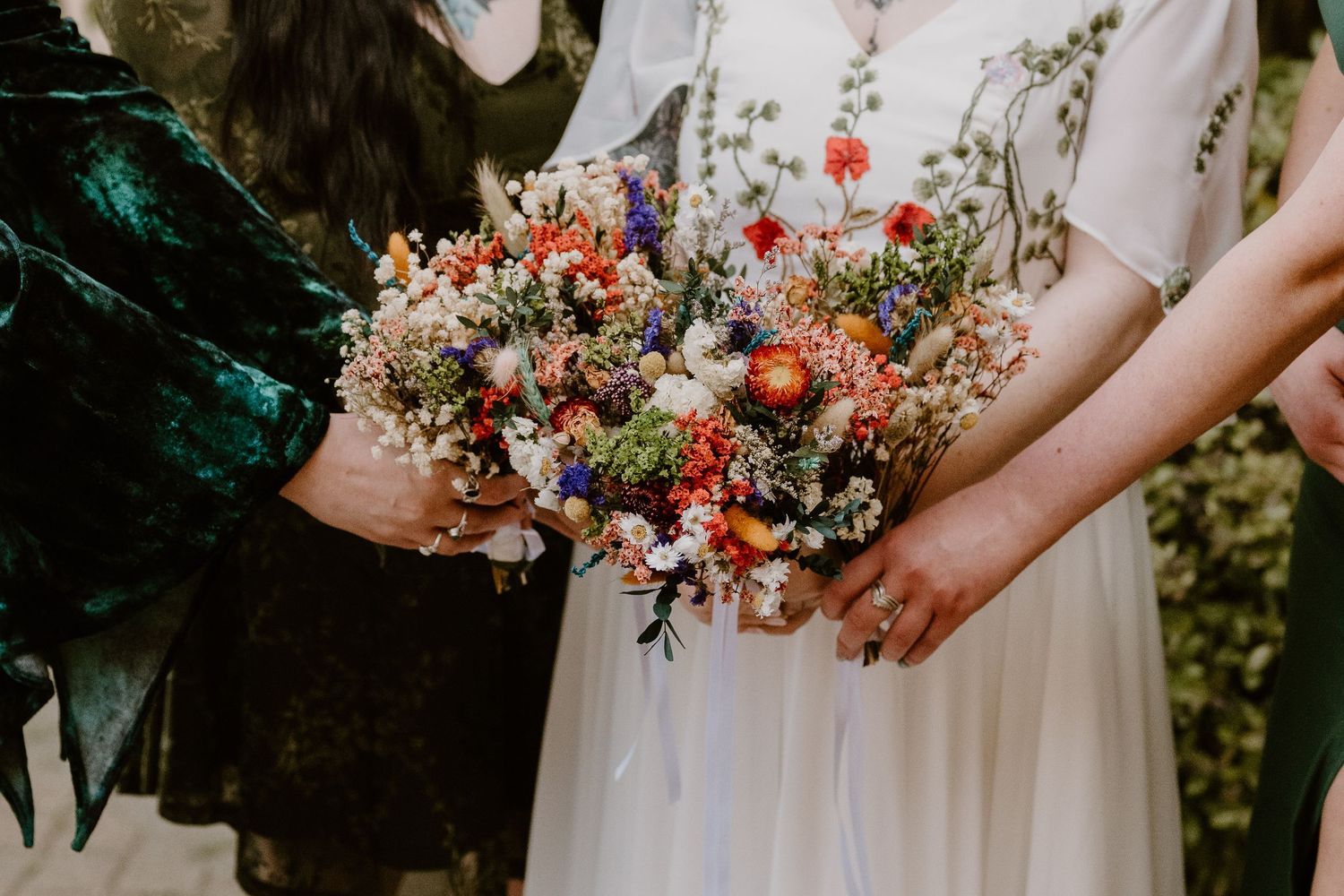 Two brides holding colorful wildflower wedding bouquets with red, purple, and white blooms outdoors.