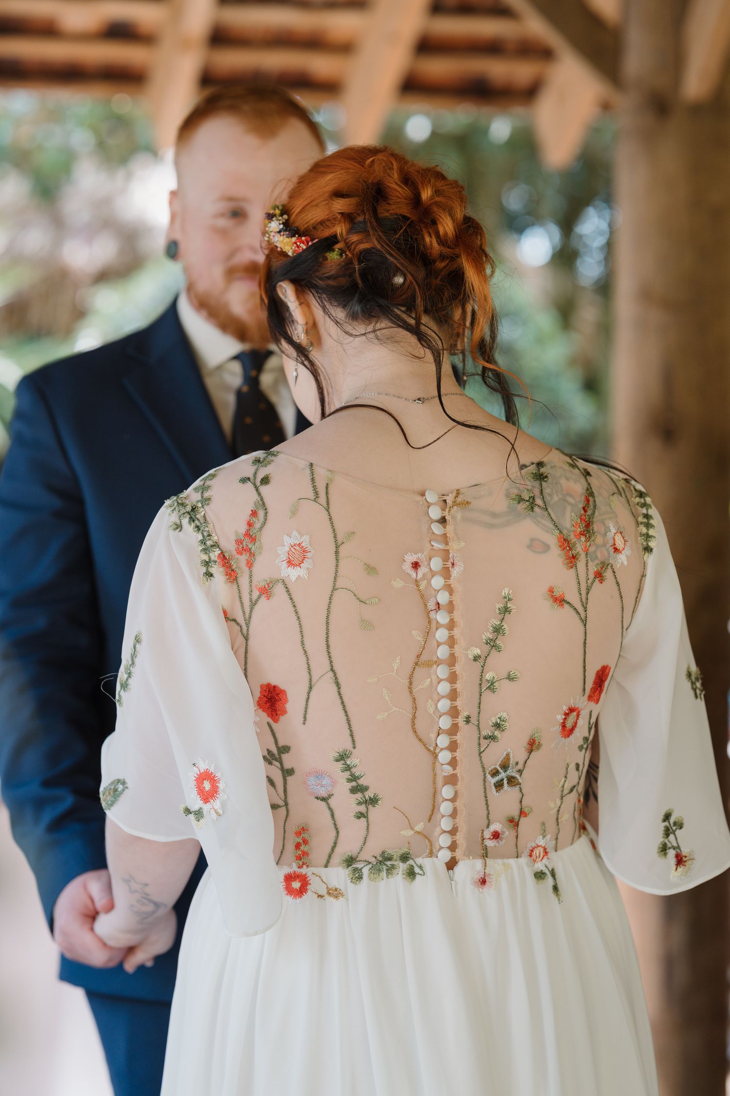 Bride in floral embroidered sheer back wedding dress with button detail, standing with groom in navy suit at rustic venue.