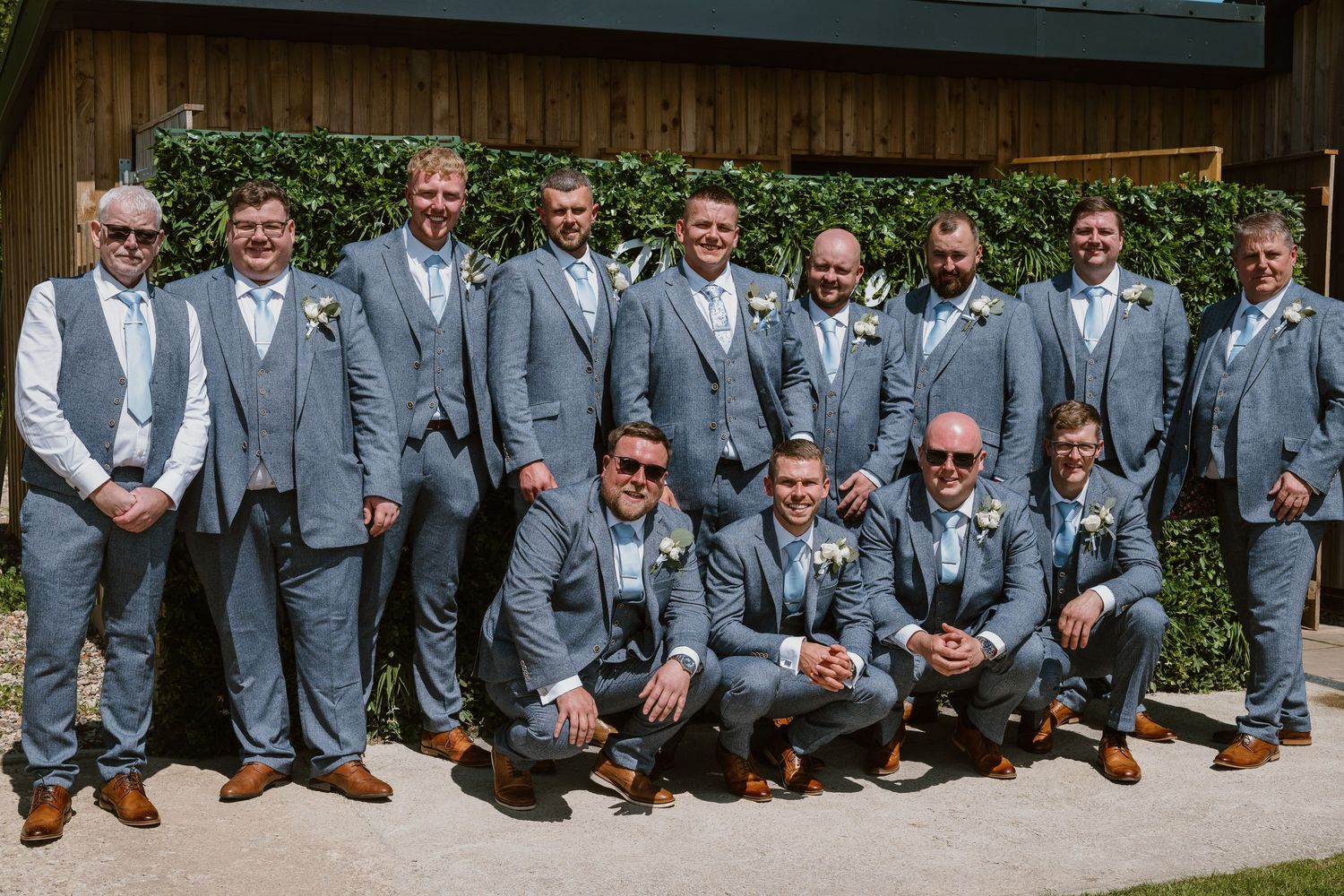 Groomsmen in matching light grey suits with brown shoes posing outdoors at a wedding venue on a sunny day.
