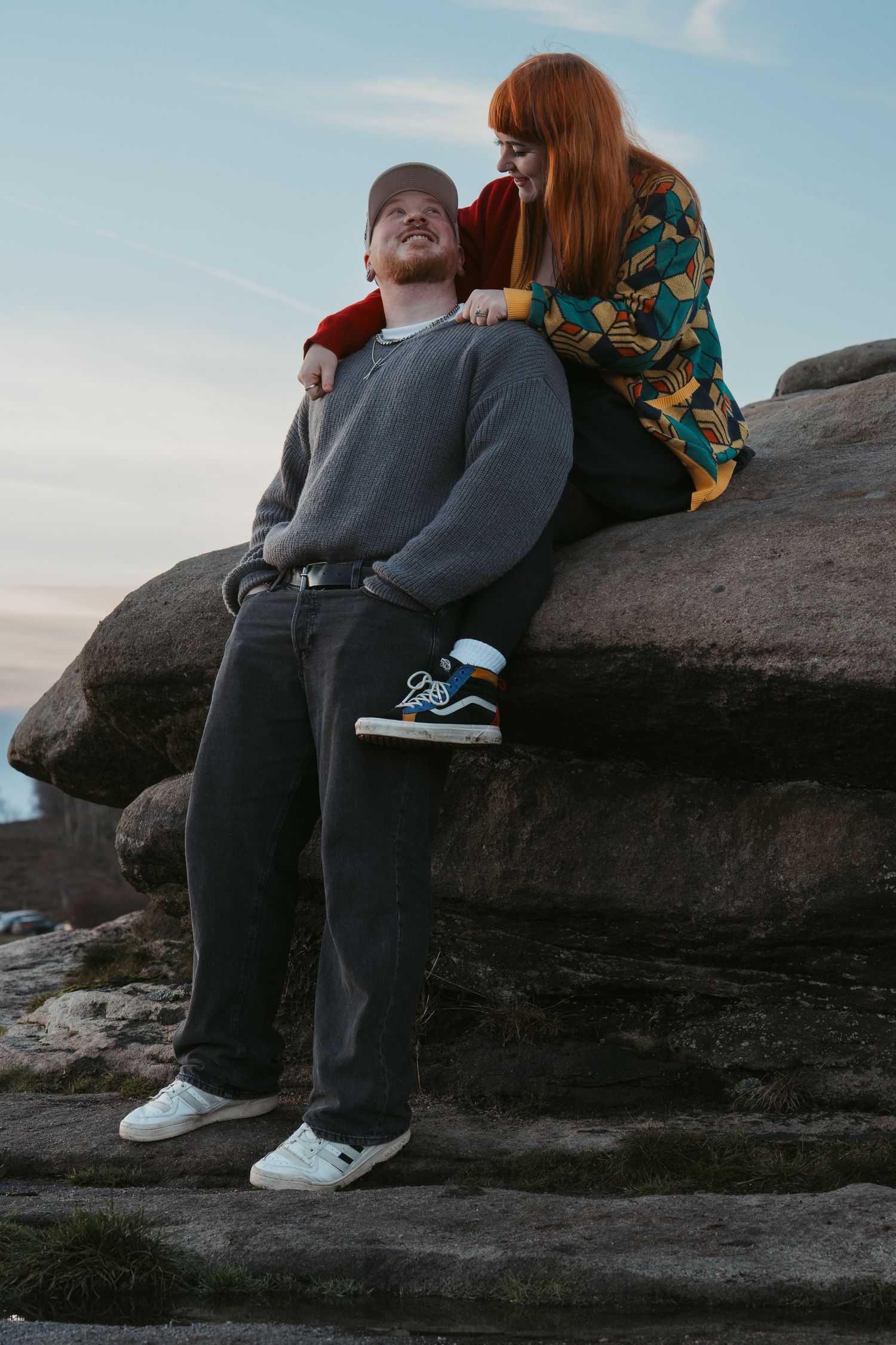 Two people sit together on a rocky outcrop at sunset wearing casual clothing and sneakers.