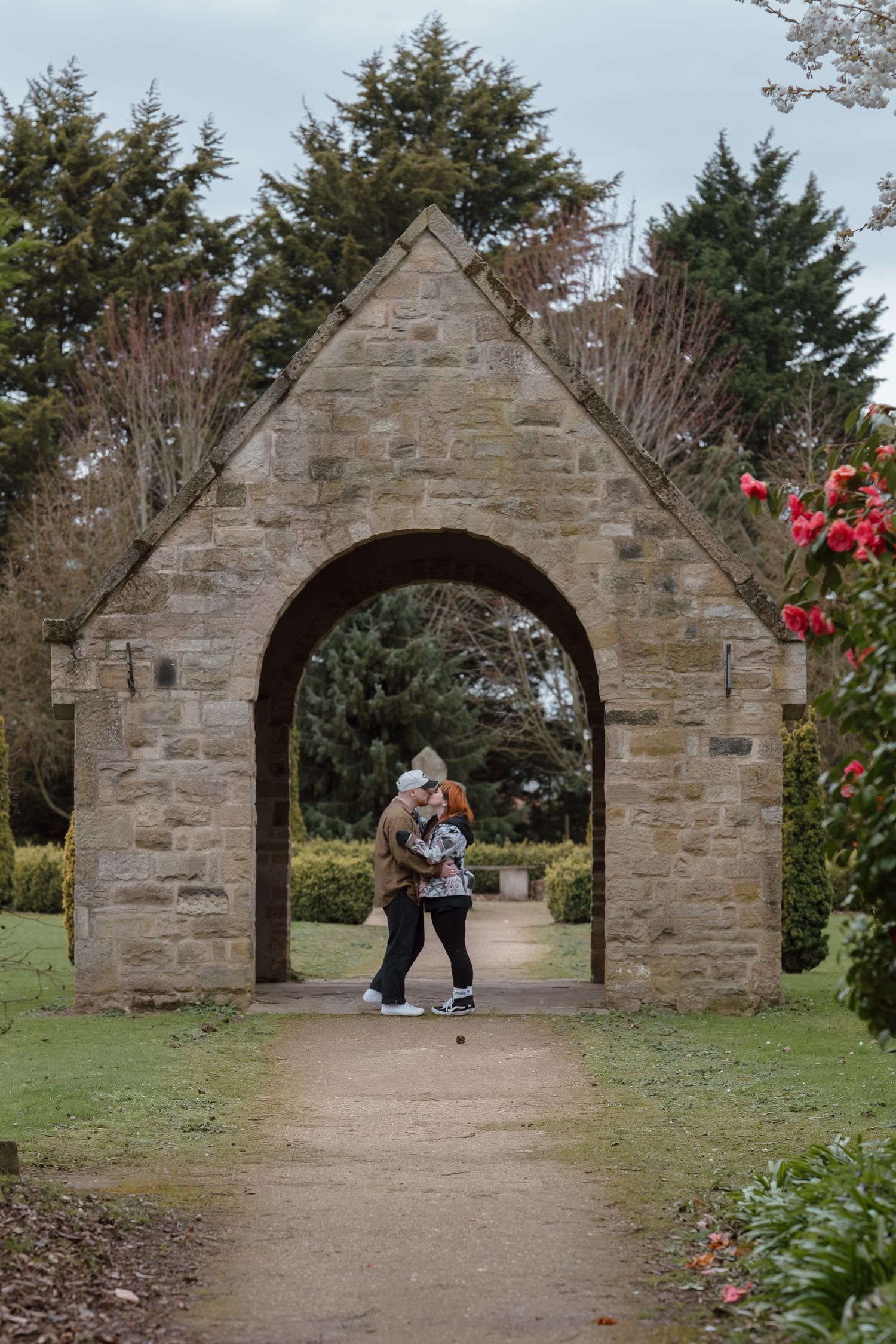 Couple embracing under a stone arch gateway in a lush garden park setting.
