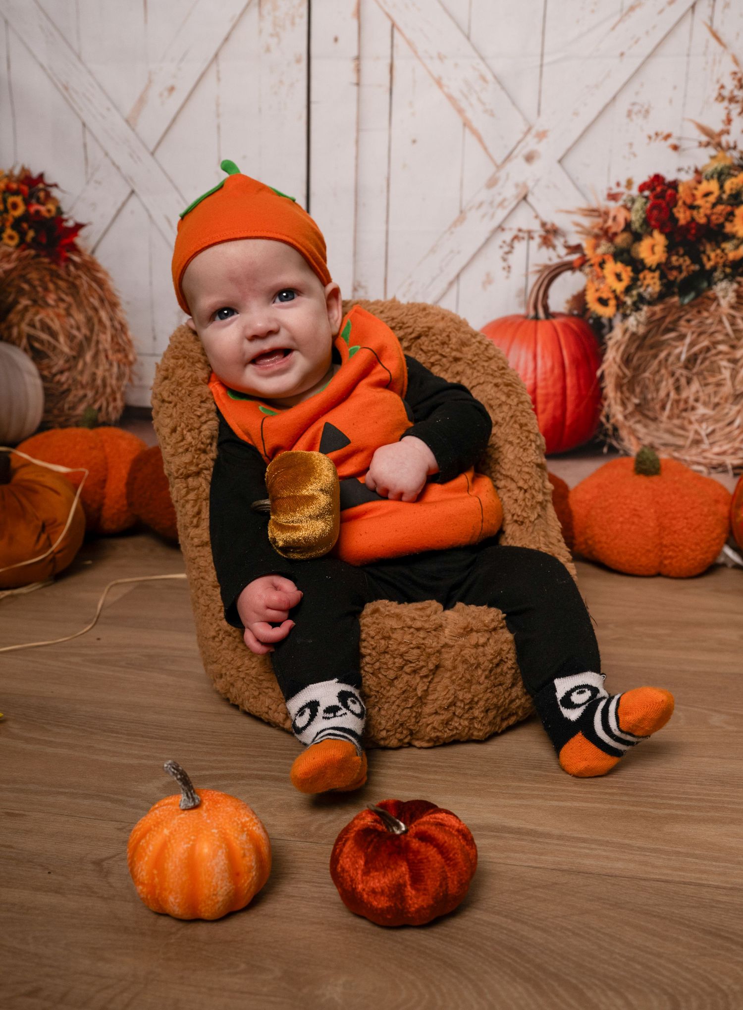 A baby in an orange pumpkin costume sits among fall decorations with hay bales and orange pumpkins on a wooden floor.