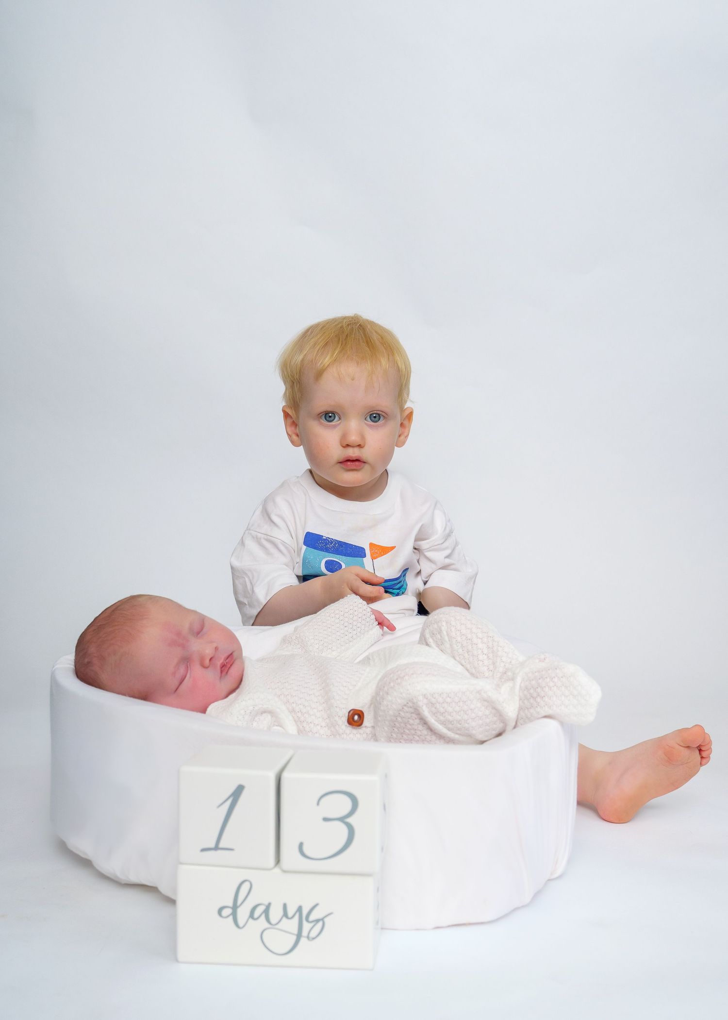 Toddler boy sits behind sleeping newborn baby at 13 days old in a white studio newborn photography session.