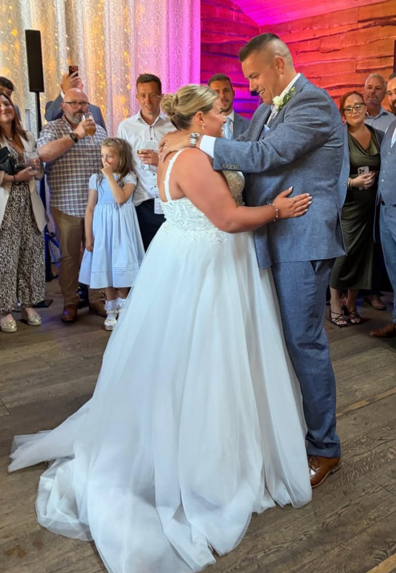 Bride and groom share their first dance at wedding reception, surrounded by guests, bride in white gown, groom in blue suit.