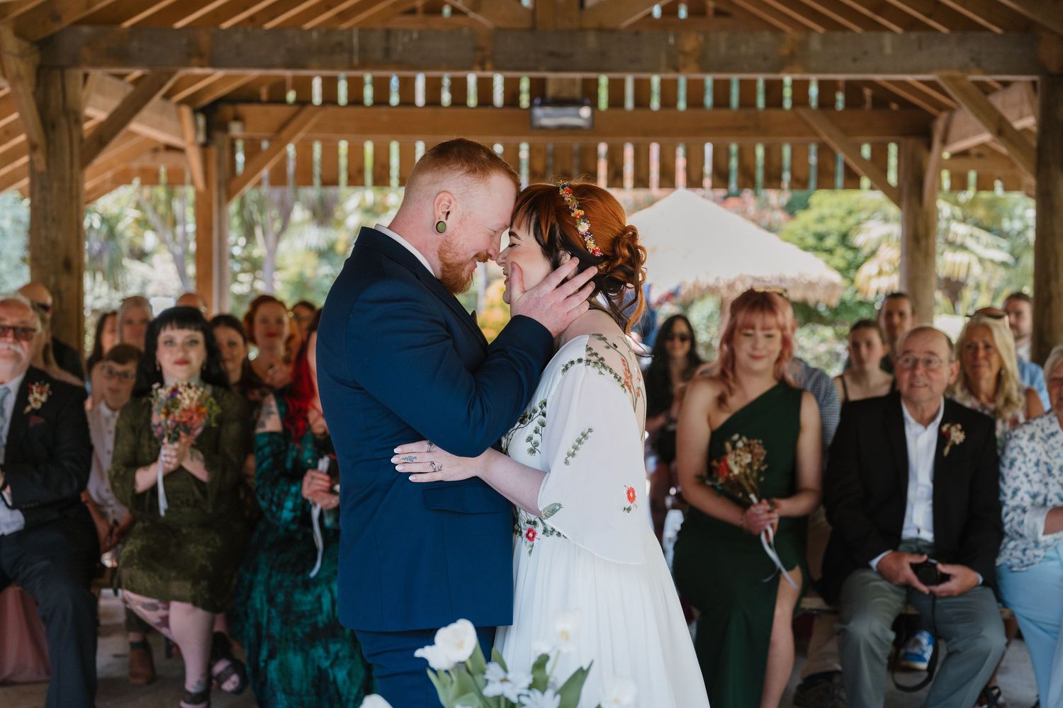 Bride and groom share their first kiss at an outdoor wedding ceremony under a wooden pavilion with guests seated behind.