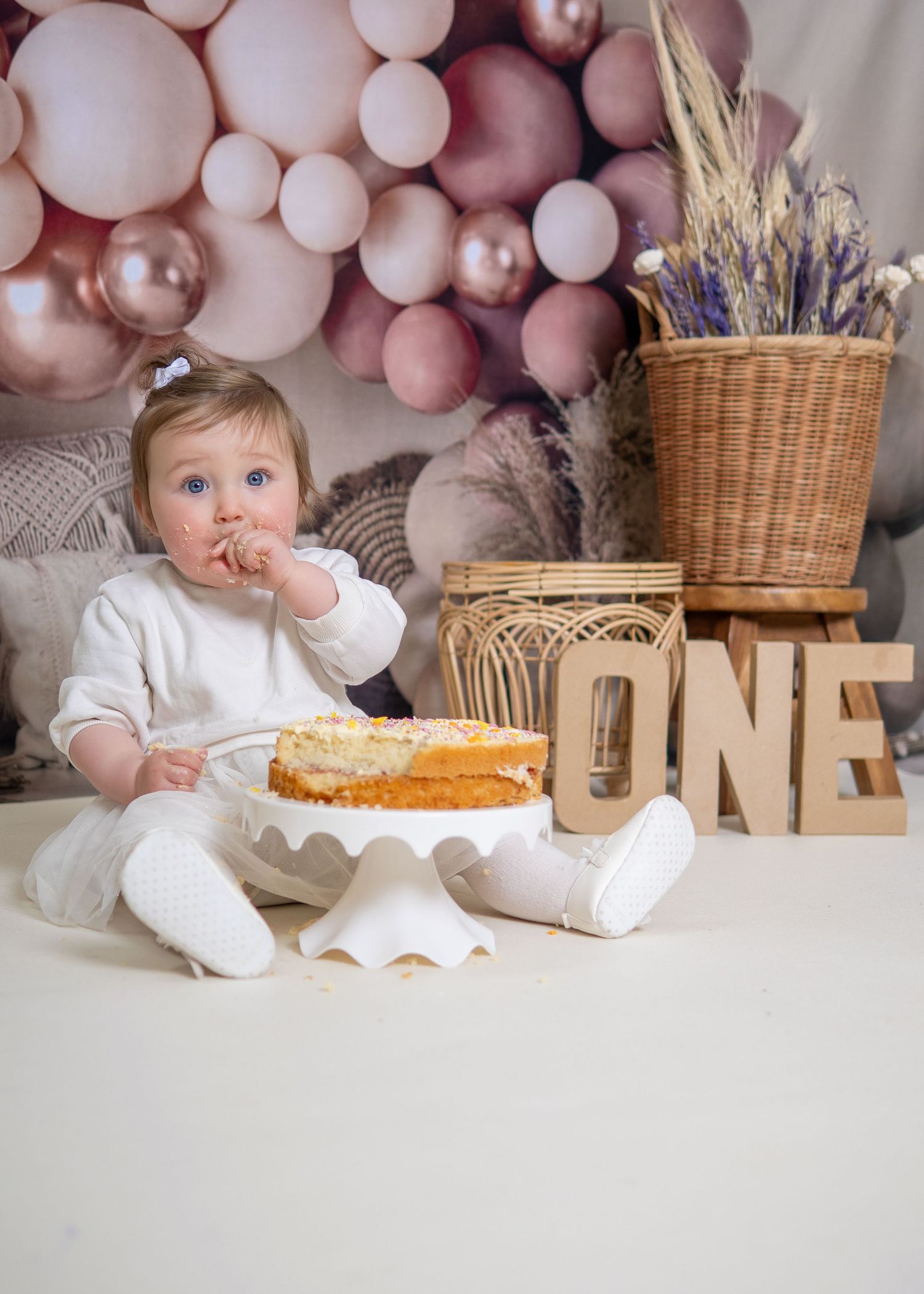 First birthday cake celebration setup with pink balloon arch, cake and rustic decor against neutral backdrop. Cake Smash