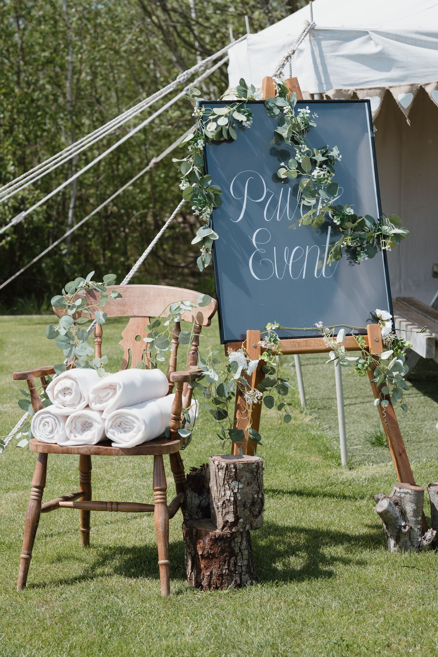 Outdoor patio event sign on easel with greenery garland, wooden chair with rolled towels, and white tent backdrop.