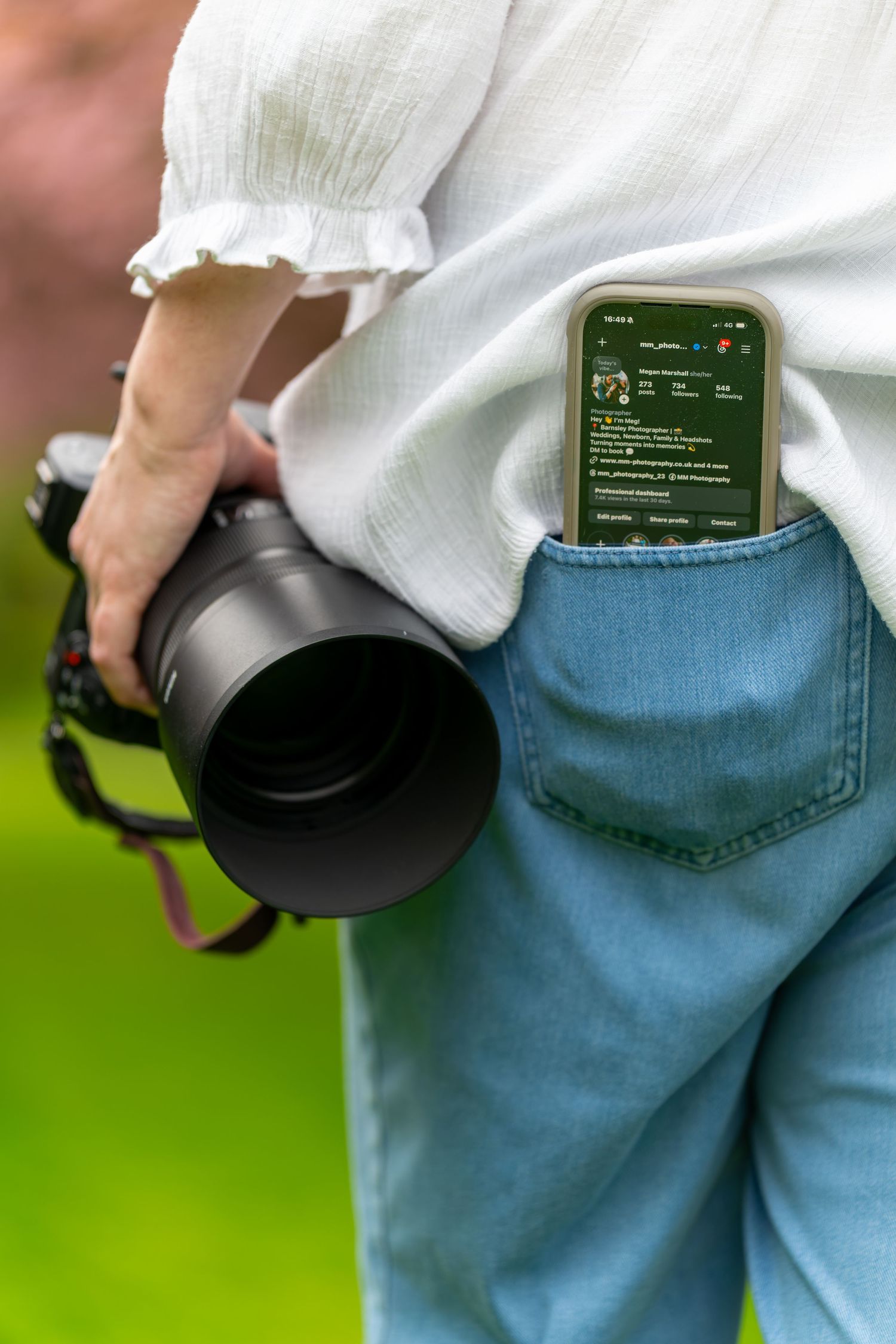 Photographer holding DSLR camera with smartphone in back pocket of jeans, outdoors with green background.