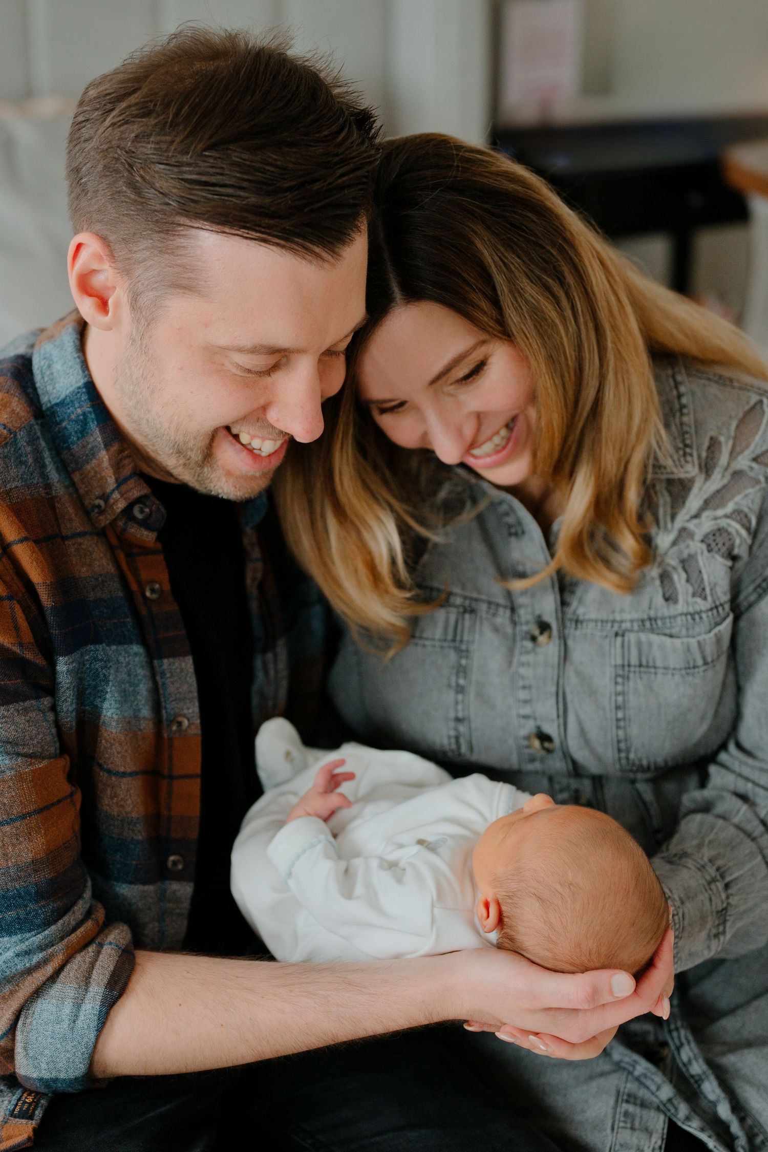 Smiling couple in casual flannel and denim jackets gently cradle a sleeping newborn baby wrapped in white, sharing a tender family moment.