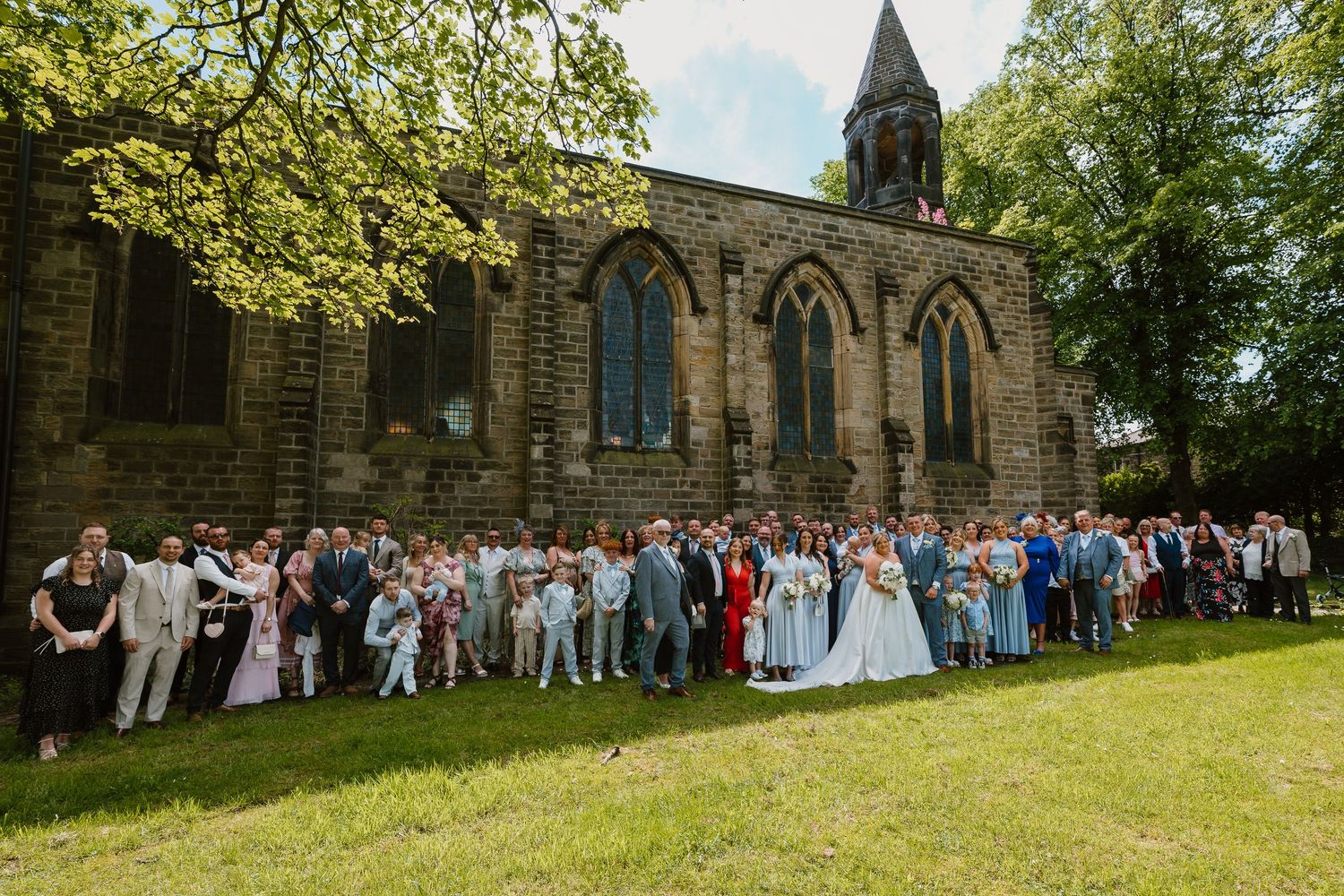 Large wedding party posed outside historic stone church on sunny day with lush green trees.