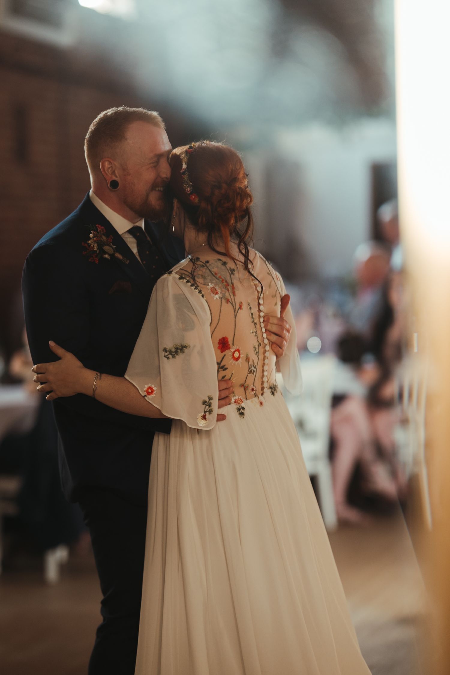 Bride and groom share first dance in warm light, bride wearing embroidered white gown with floral details at elegant wedding reception.