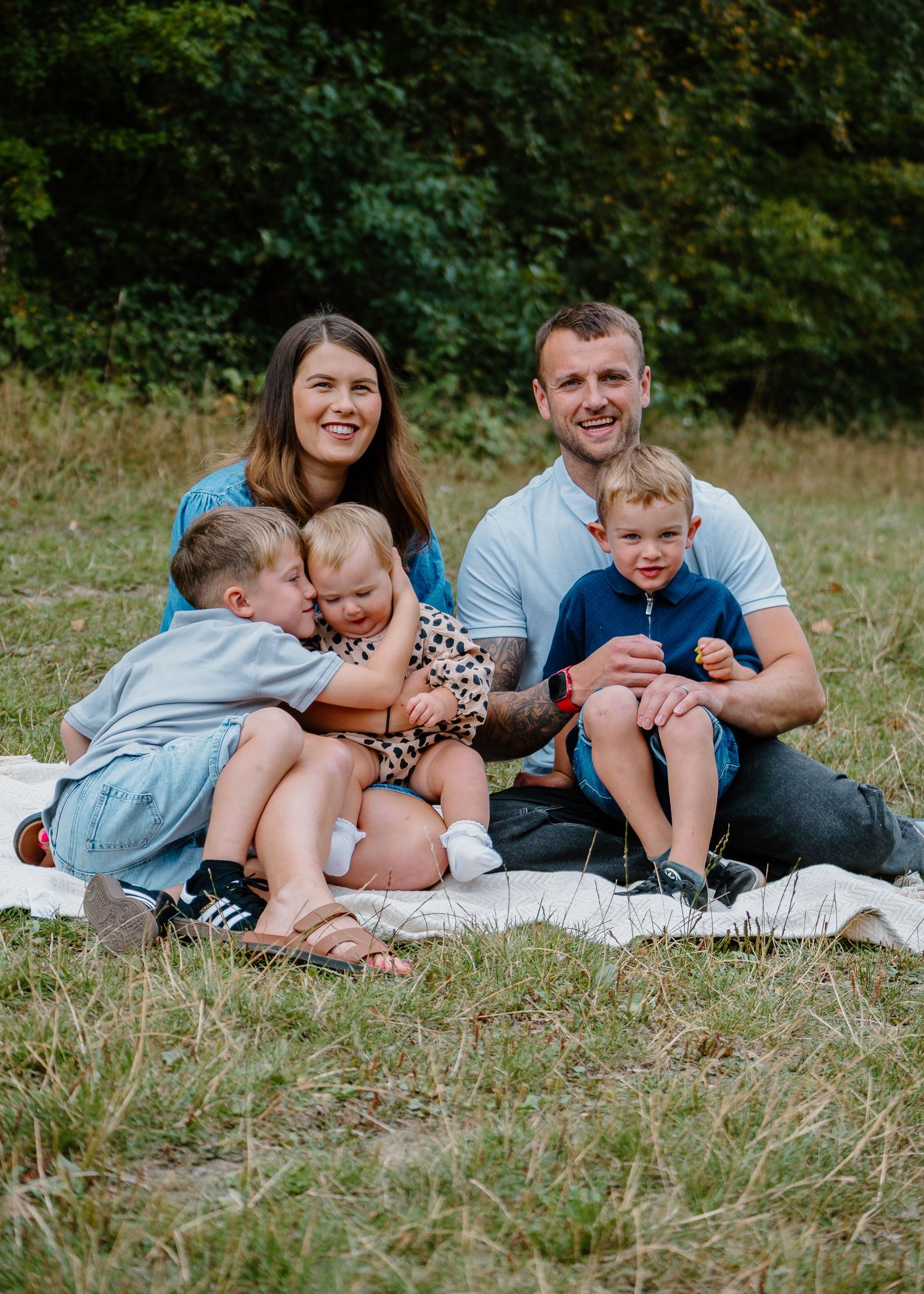Family Photoshoot - A family sits together on a blanket outdoors during a summer photo session in a grassy field with trees in background.