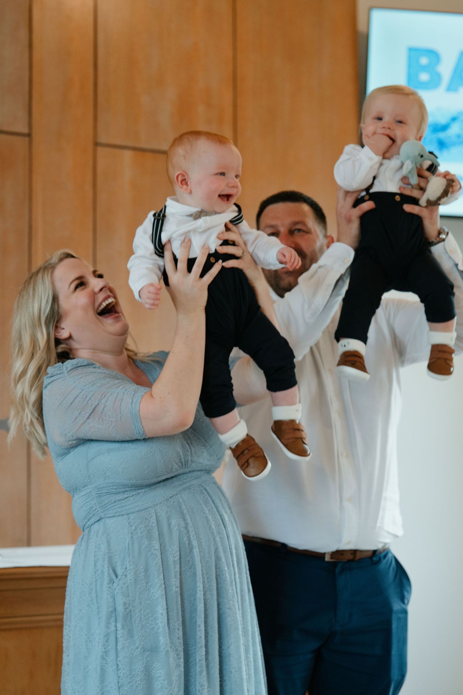 A joyful couple laughs while lifting two babies dressed in matching black and white outfits during a celebration.
