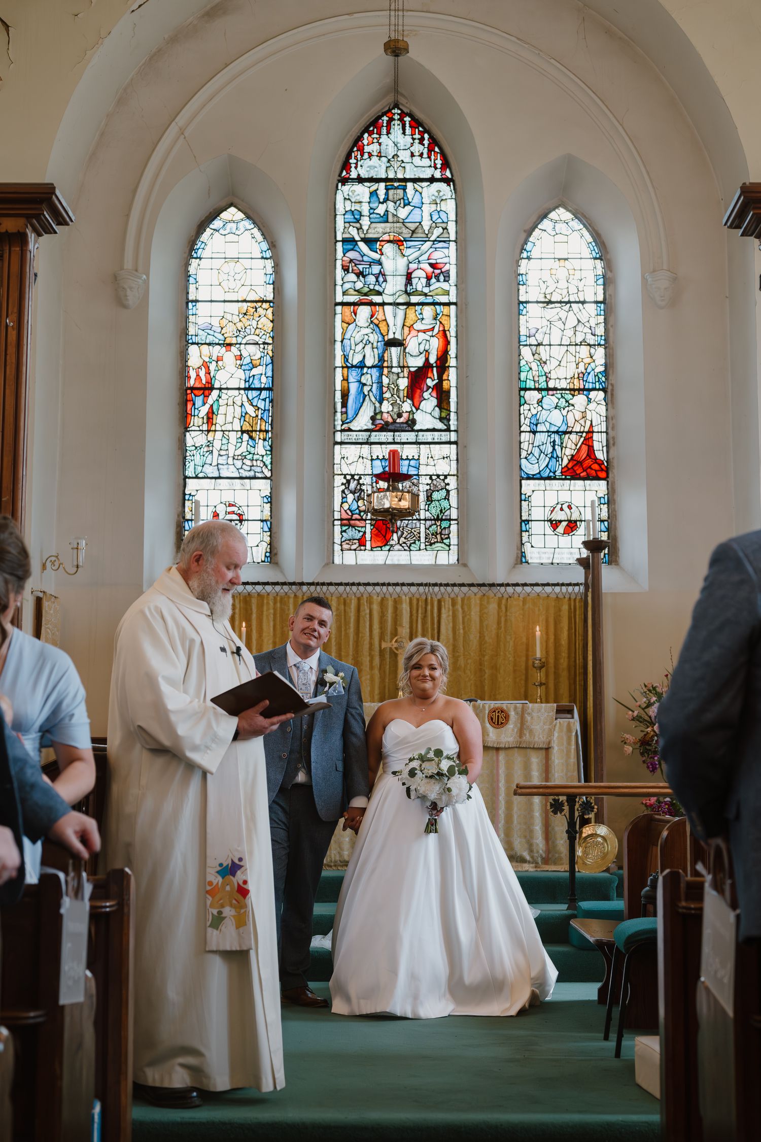 Bride and groom exchange vows at church altar with stained glass windows and officiant holding ceremony book.