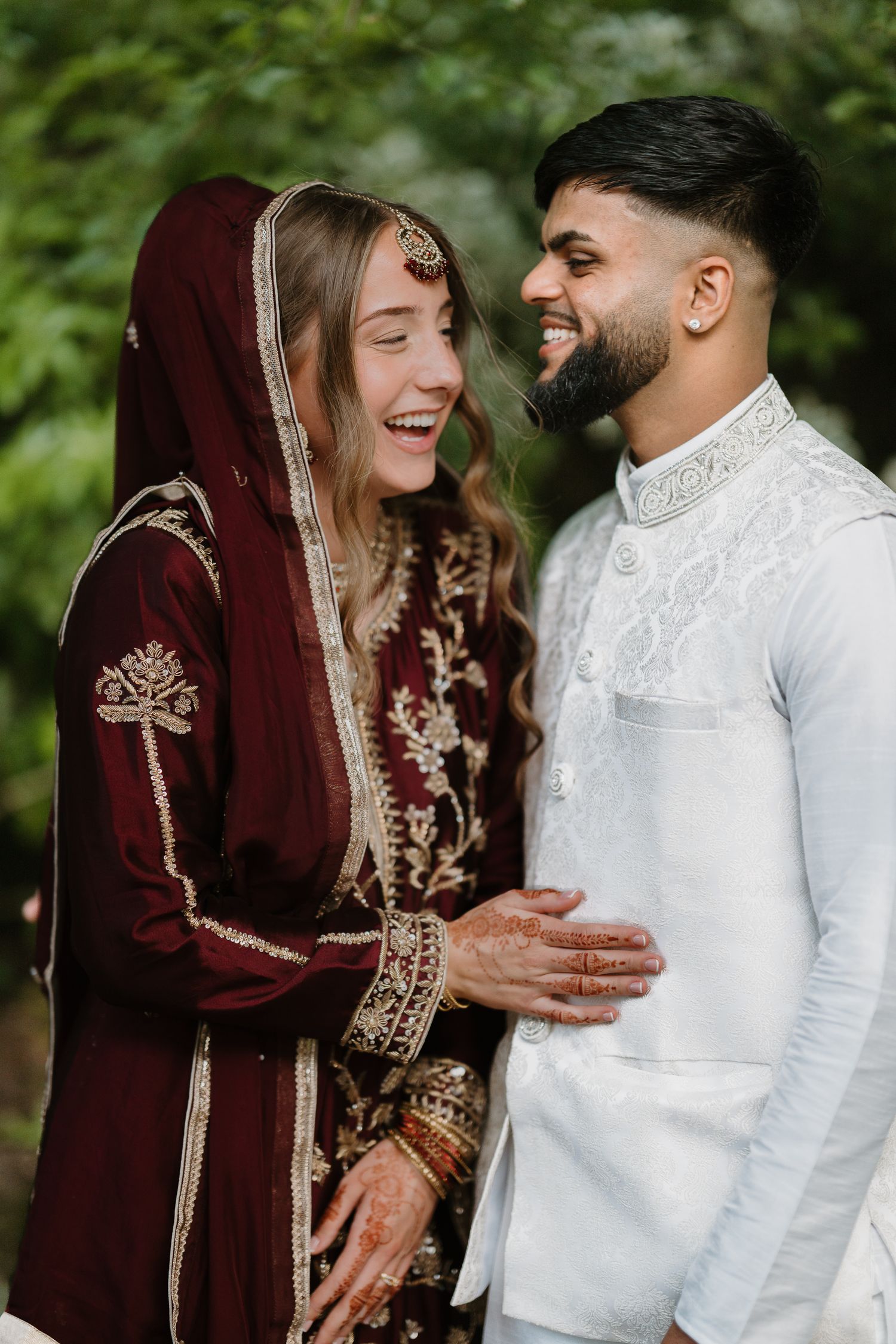 A joyful couple in traditional South Asian wedding attire, sharing a laugh outdoors, bride in maroon and groom in white.