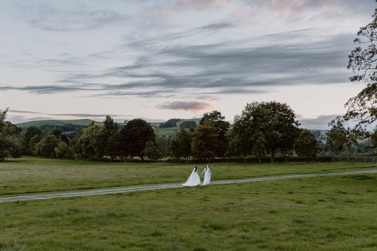 A bride and groom walk hand in hand along a path through lush green fields at sunset.