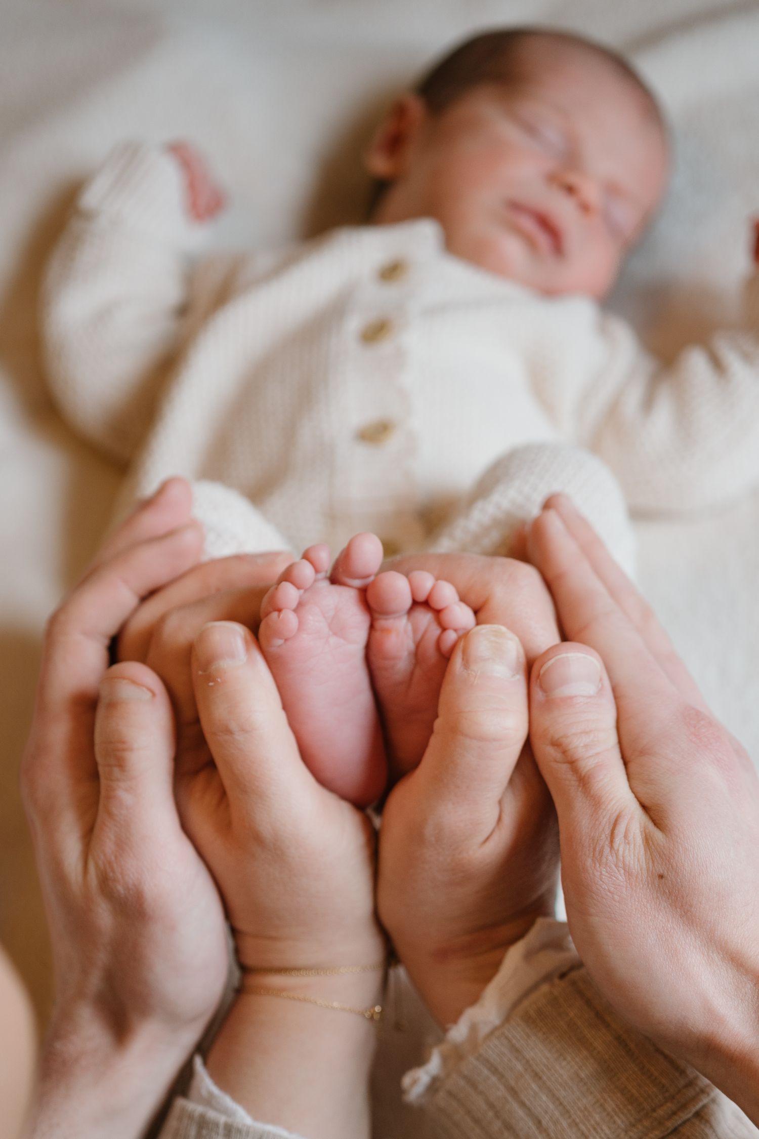 Parents cradling tiny newborn baby feet in their hands, with sleeping infant in white knit outfit in soft background.