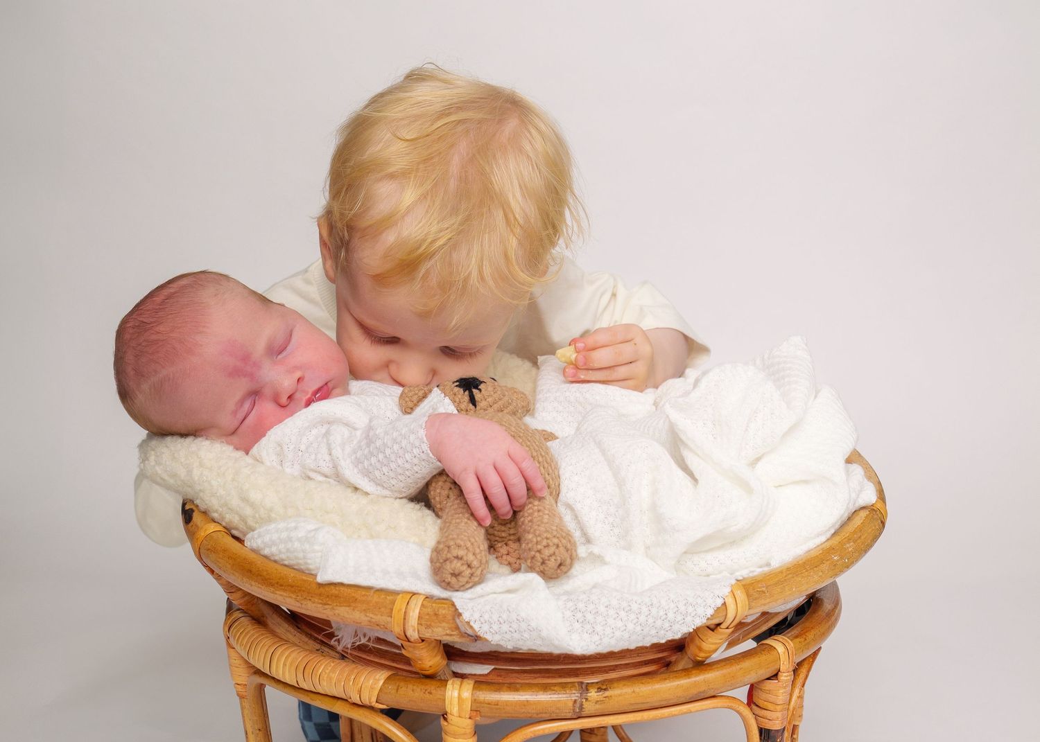 Newborn Photography - A baby in white clothing lies in a wicker basket while a young child leans in to give a gentle kiss.