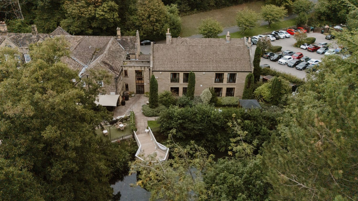 Aerial view of a historic stone manor house surrounded by lush trees with a car park visible in the background.