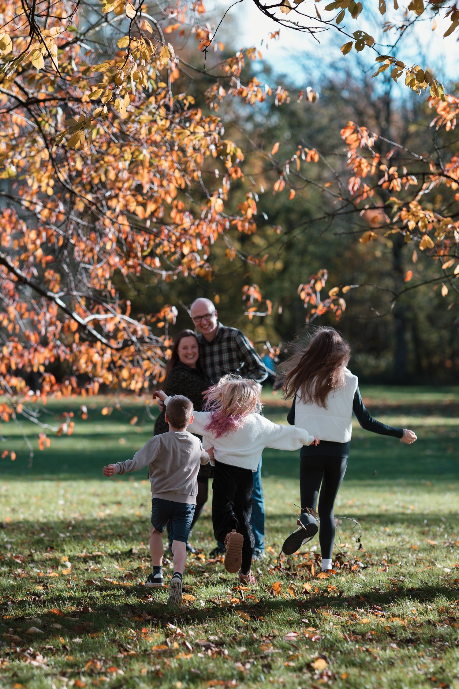 A family walks together through a park on an autumn day with golden leaves falling from trees in the background.