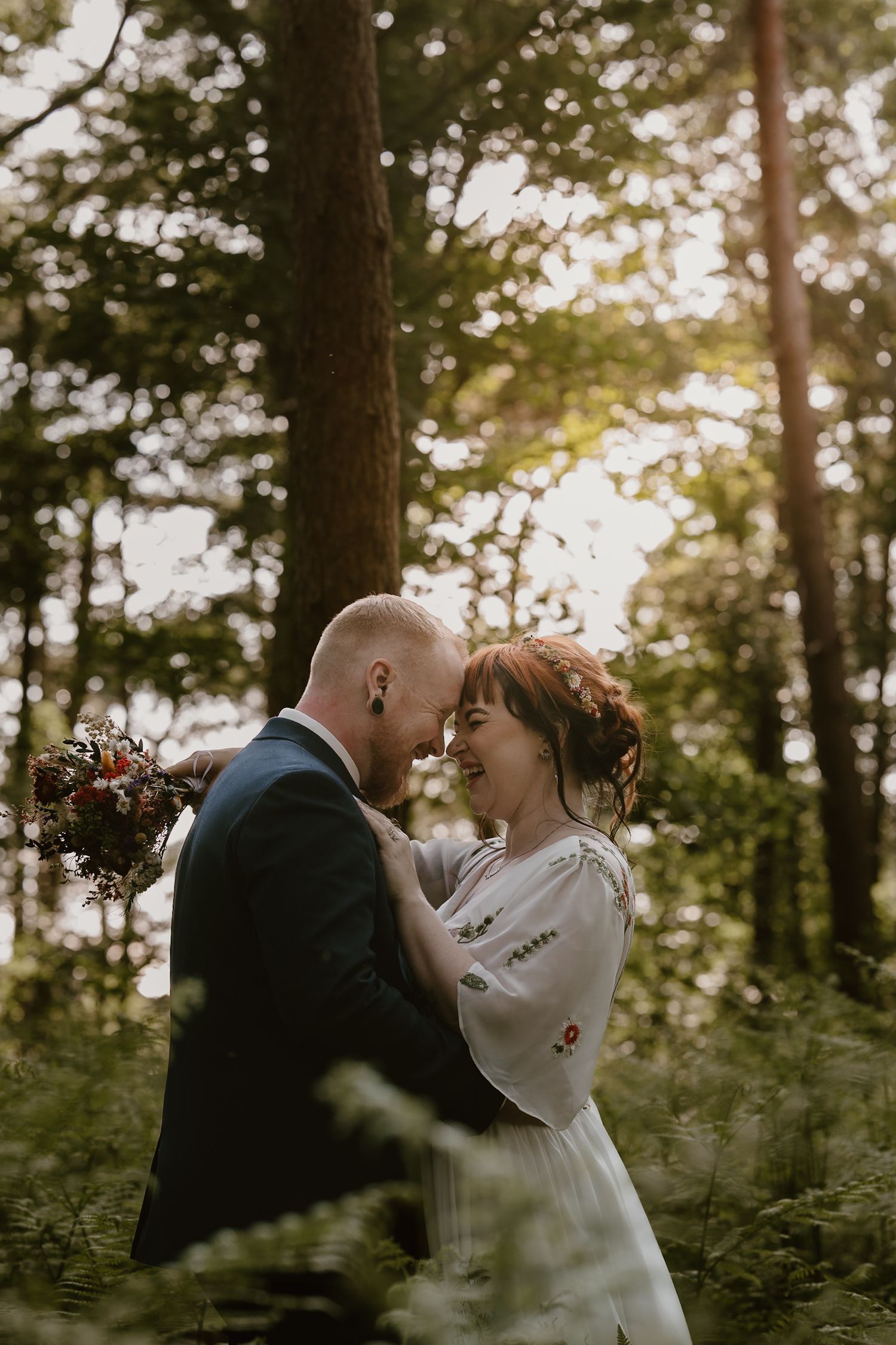 A bride and groom share a romantic moment in a lush forest, smiling at each other, with the bride holding a bouquet.