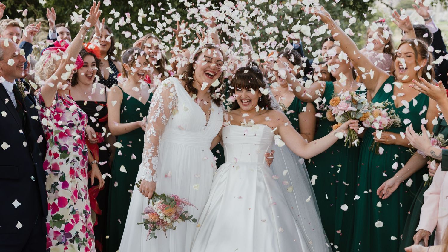 Two brides in white wedding gowns celebrate with guests tossing white confetti petals outdoors.