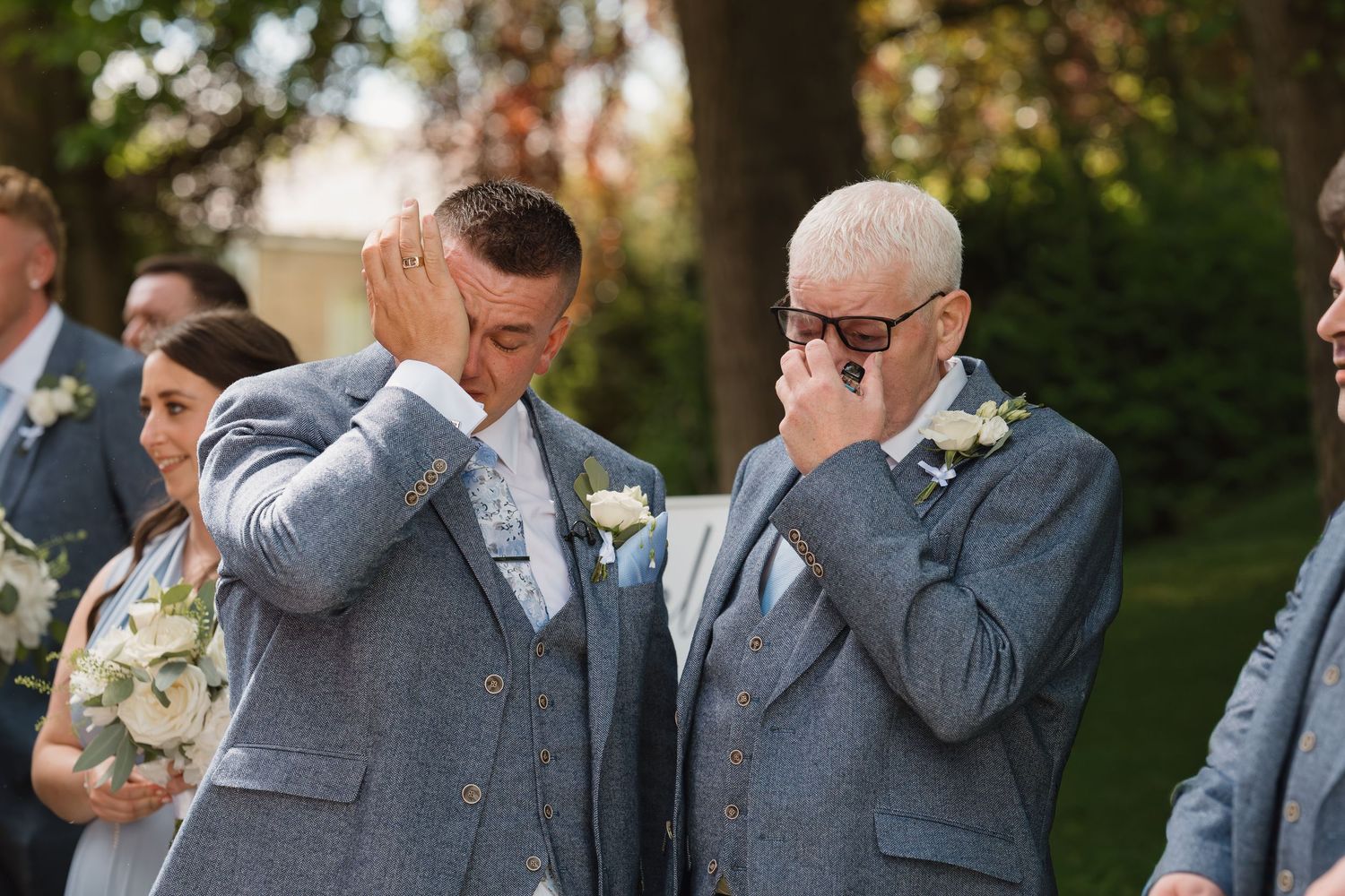 Two emotional grooms in matching grey suits wipe tears at their outdoor wedding ceremony, with groomsmen standing nearby.