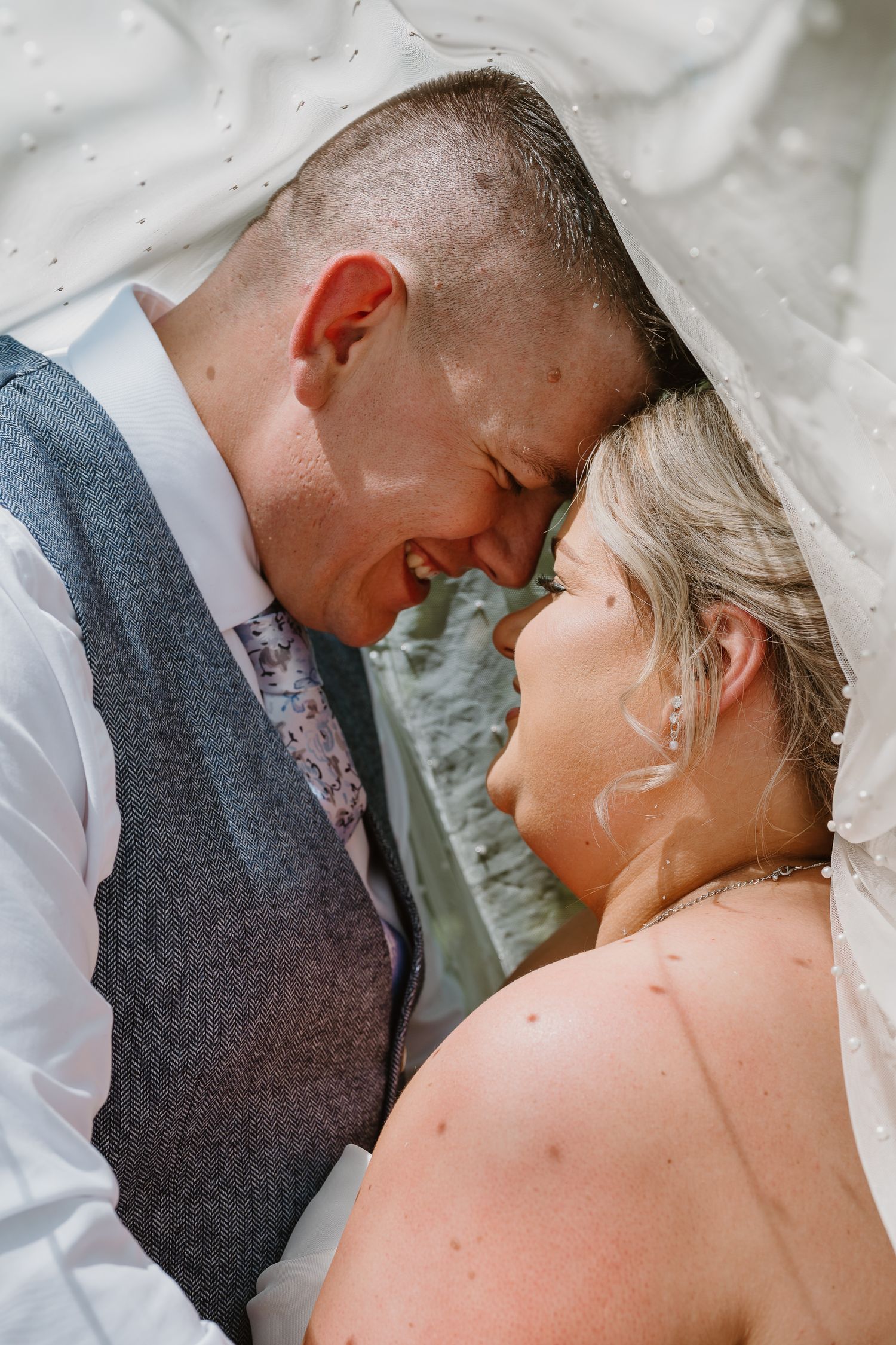 A bride and groom share an intimate moment under a wedding veil, smiling and touching foreheads on their wedding day.