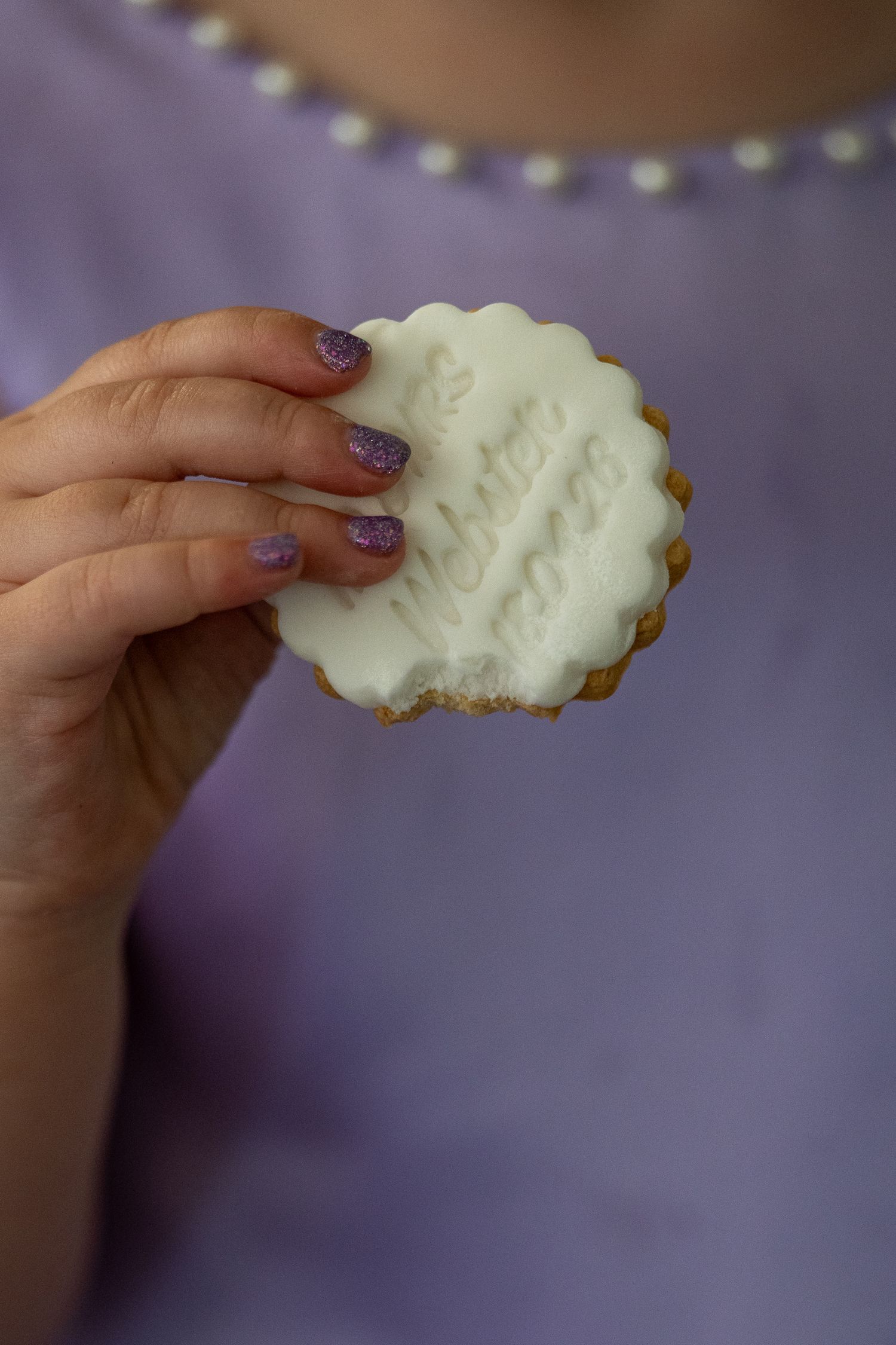 Hand with purple glitter nails holding a round white iced cookie with decorative text, against a lavender background.