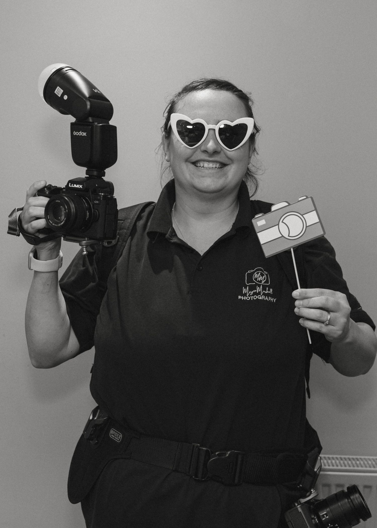 Woman photographer smiling in heart sunglasses holding Canon camera with flash and Instagram prop sign, black and white photo.