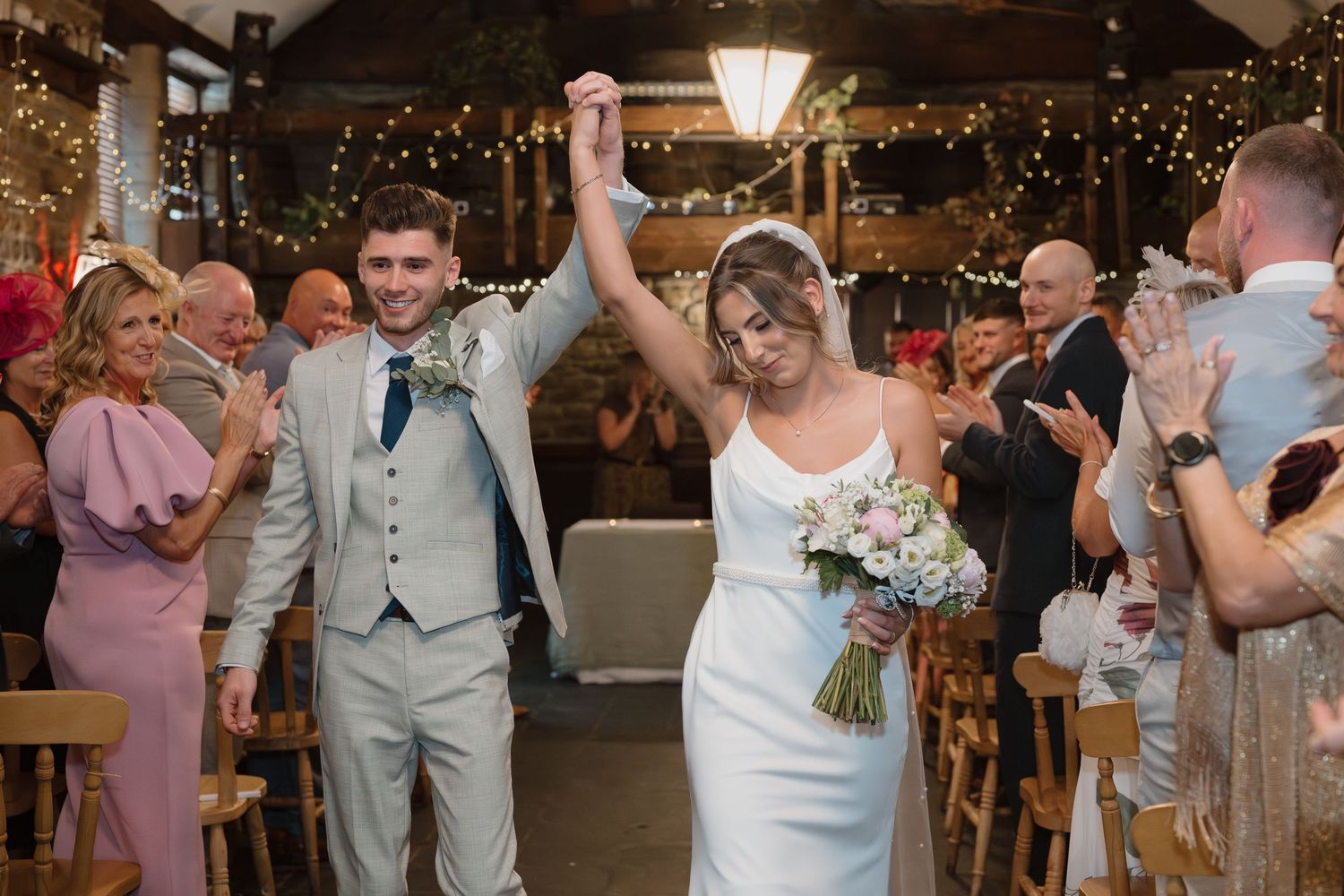 Newlywed couple celebrating their wedding recessional in a rustic barn venue with fairy lights and cheering guests.