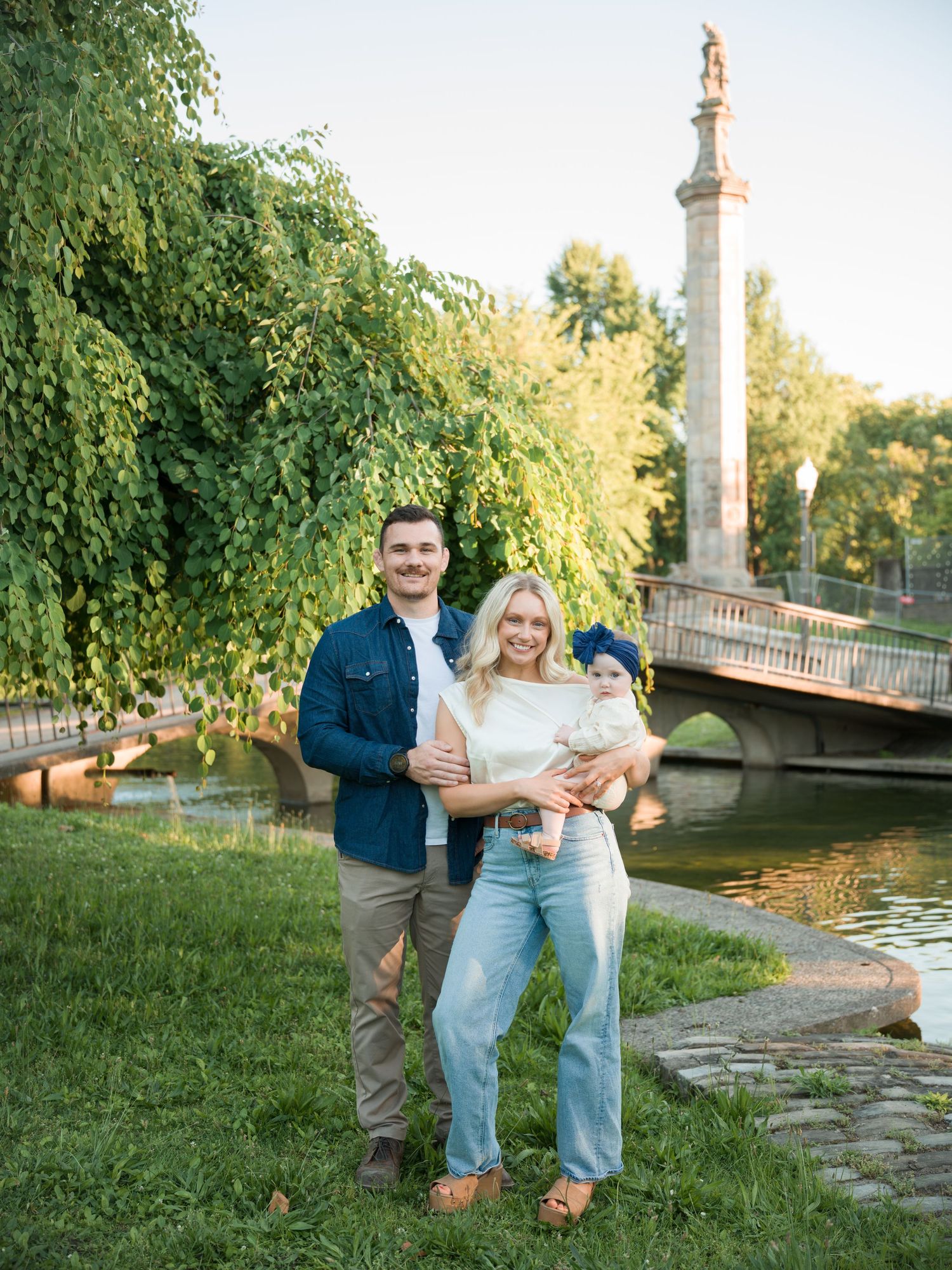 A young family of three poses outdoors near a bridge and pond, with lush green trees in golden hour light.