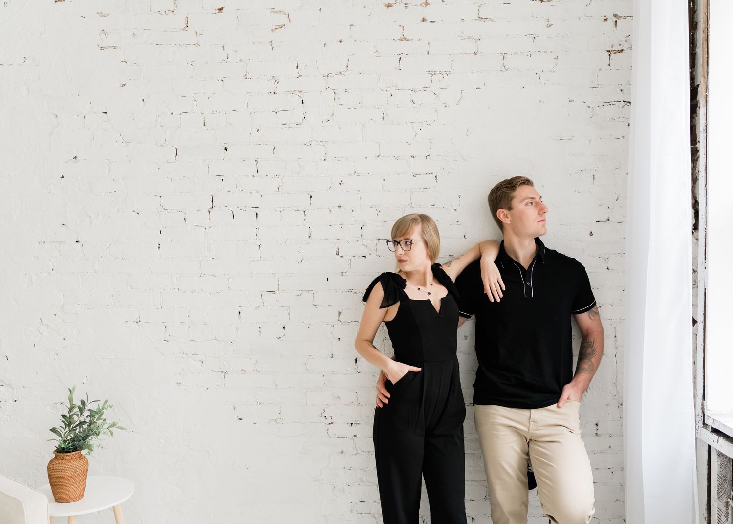 Couple in matching black outfits posing against white brick wall with minimalist decor.