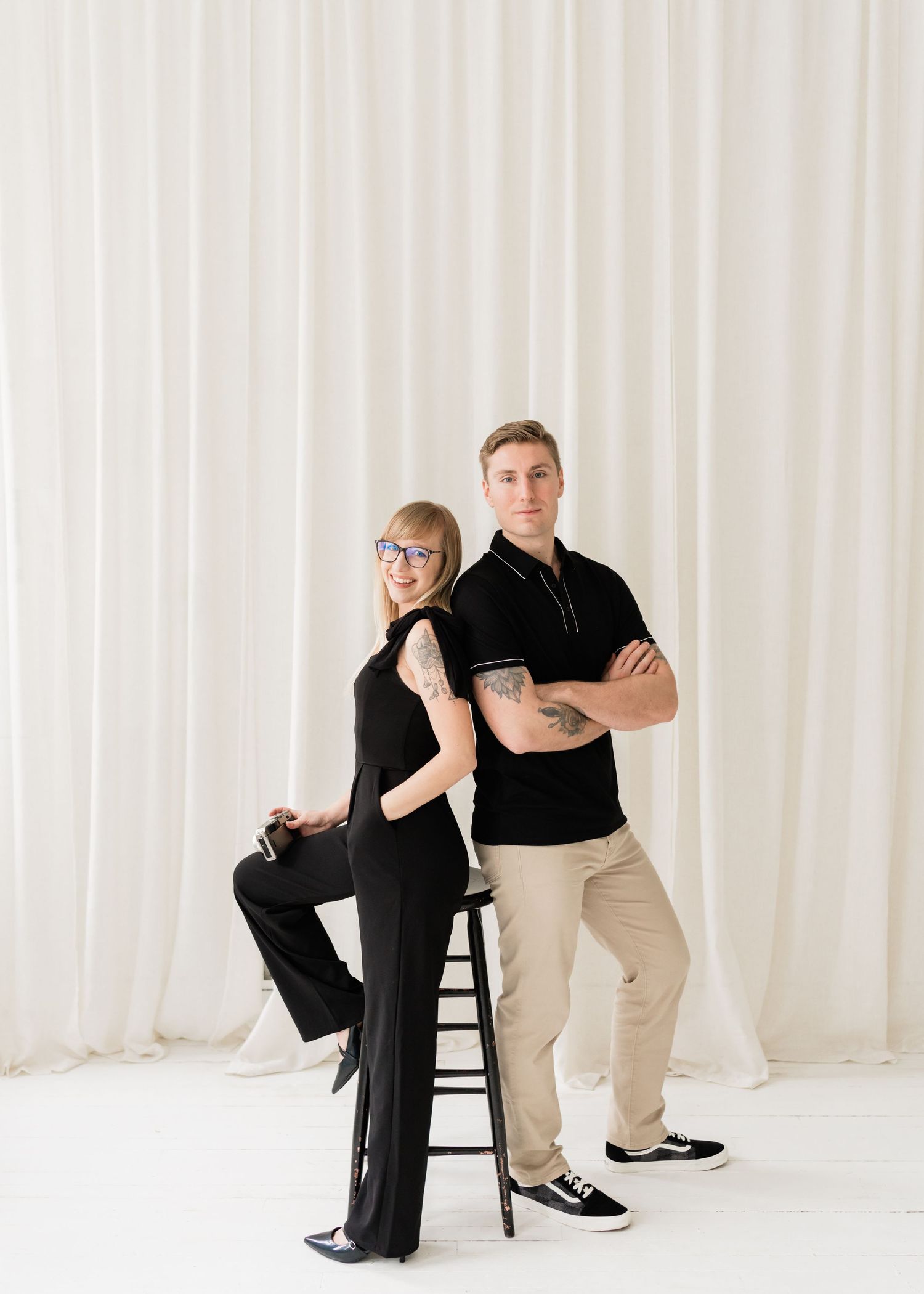 Studio portrait of couple in black and khaki outfits posing against white curtain backdrop.