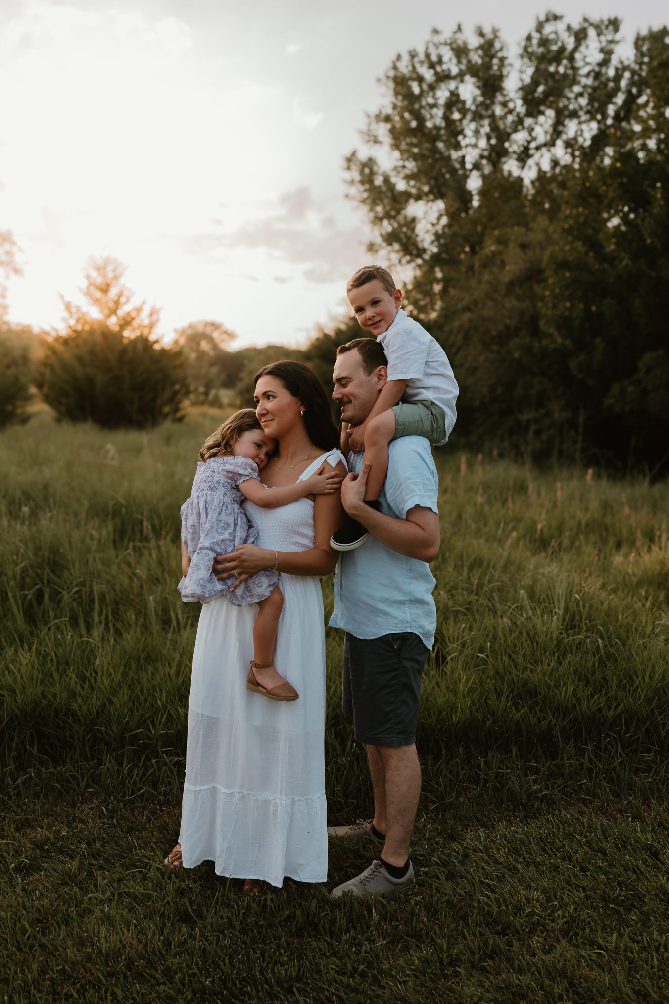 A family shares a tender moment together in a grassy field at sunset, with trees silhouetted in the background.