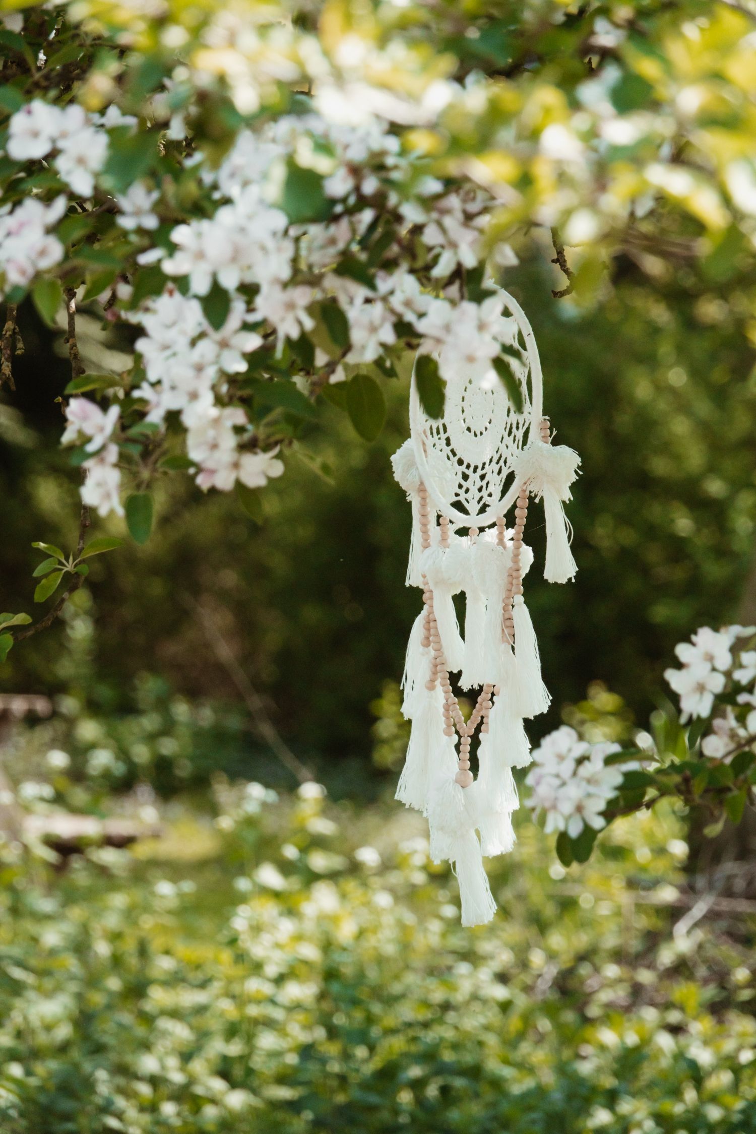 A white boho dreamcatcher hangs from a flowering apple tree branch in a sunny green garden.