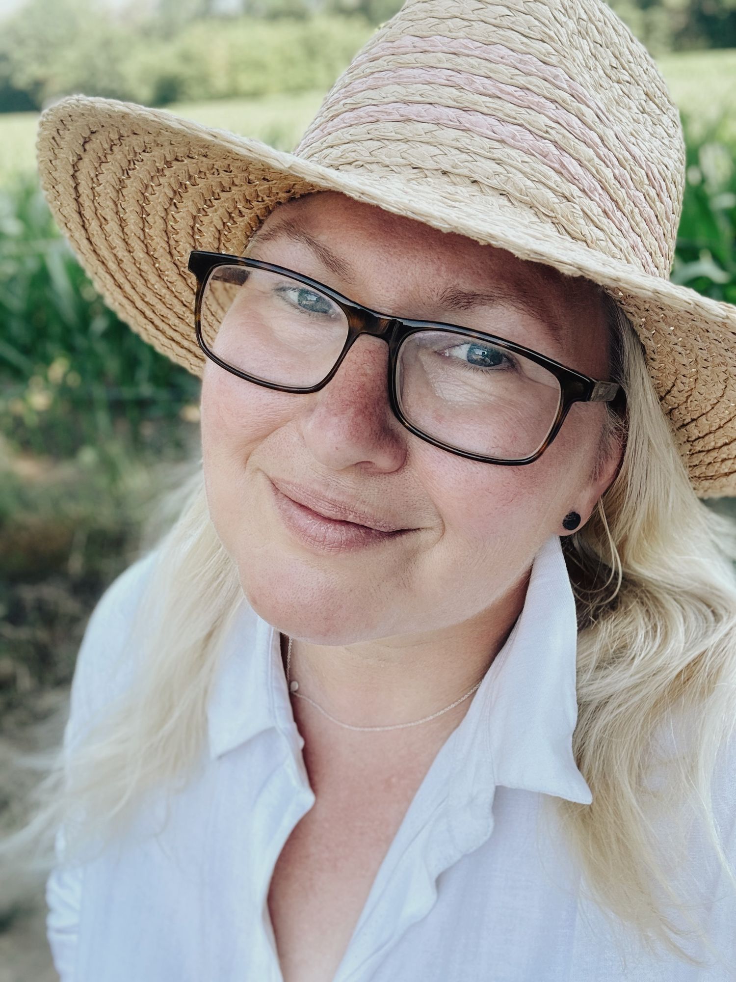 Marjolein wearing straw hat and black glasses posing outdoors near green cornfield.