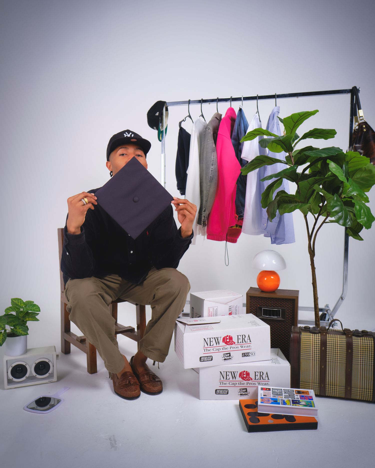 Graduate examines cap while sitting among clothing rack and study materials.