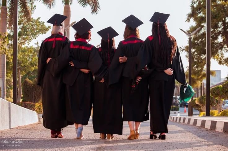 Five graduates walking together in black caps and gowns on palm tree-lined path.