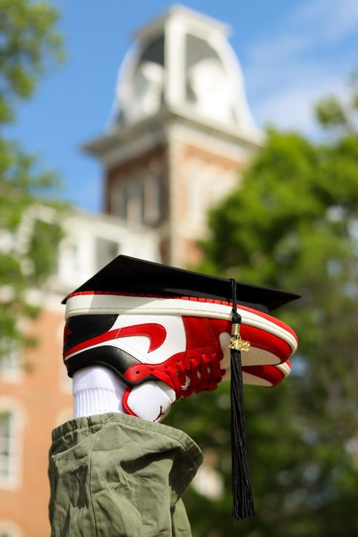 Red Nike sneaker wearing graduation cap against backdrop of classic university architecture.