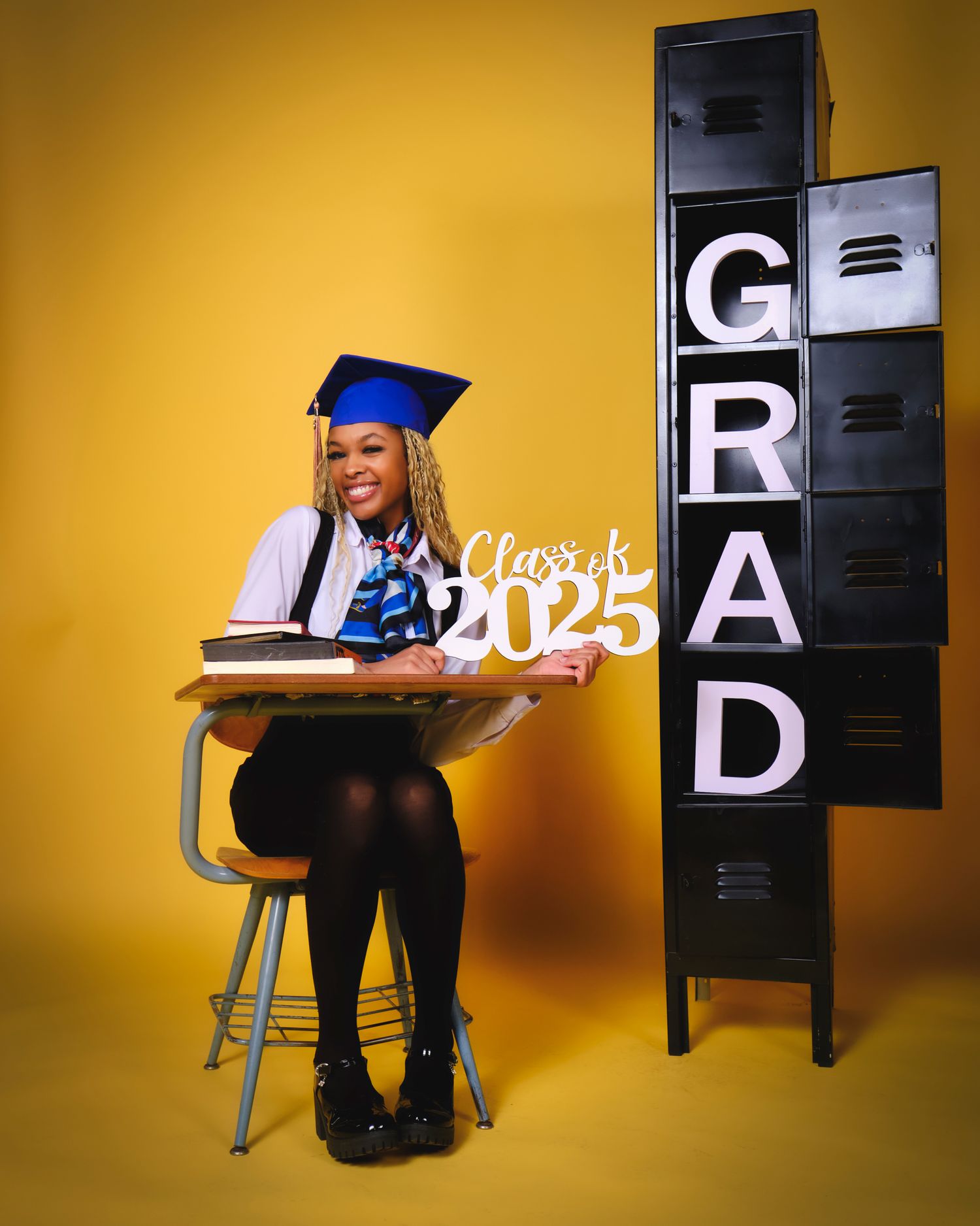 Graduate sits in school chair next to lockers with Class of 2025 text on yellow background.