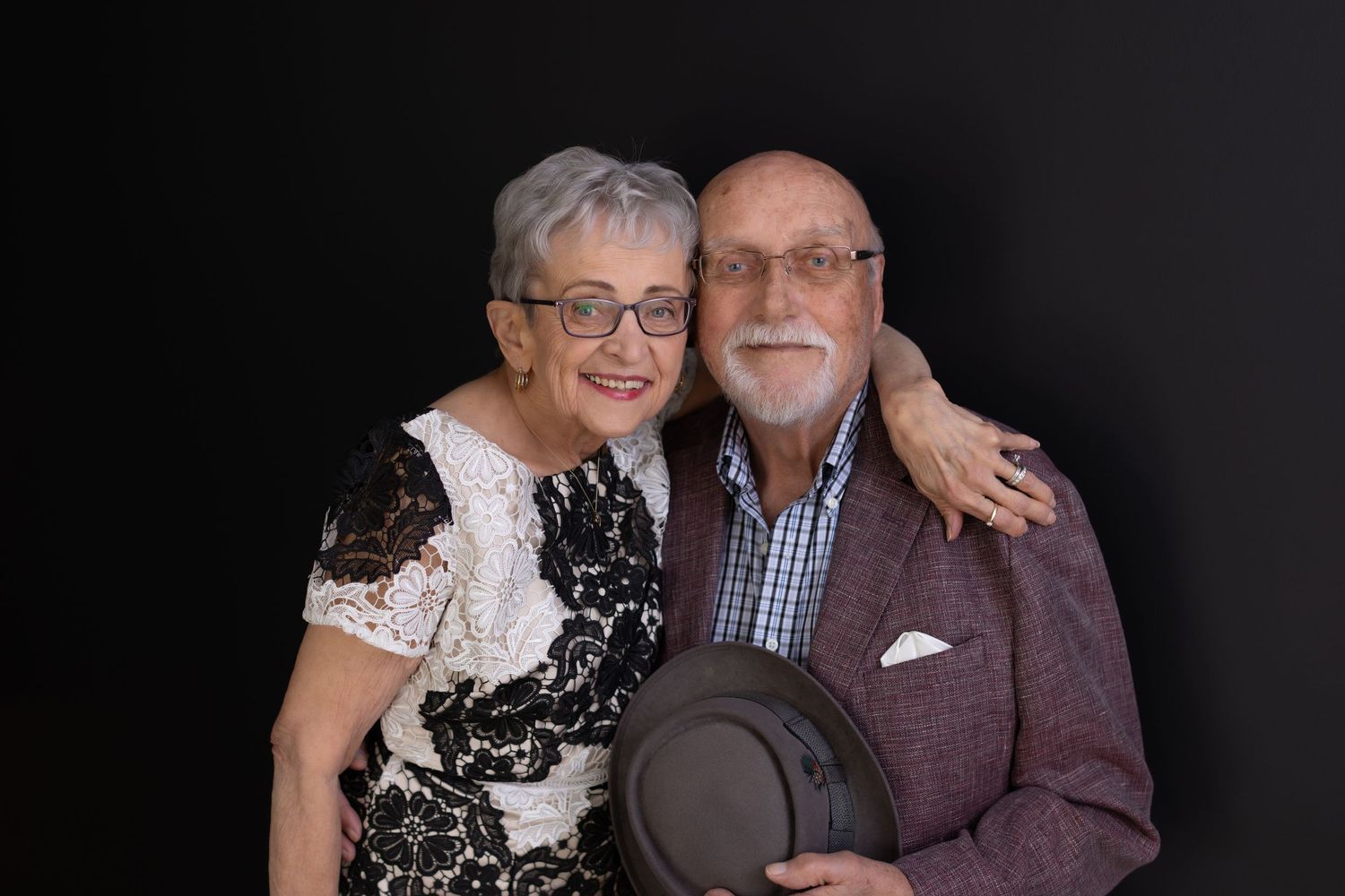Elderly couple in formal attire pose together against dark background for a portrait photo.