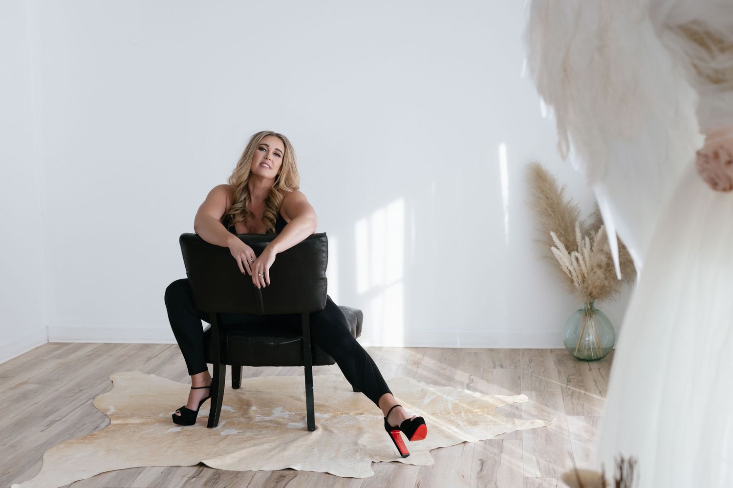 A person in black attire sits casually in a modern velvet chair against a white wall with minimalist decor.