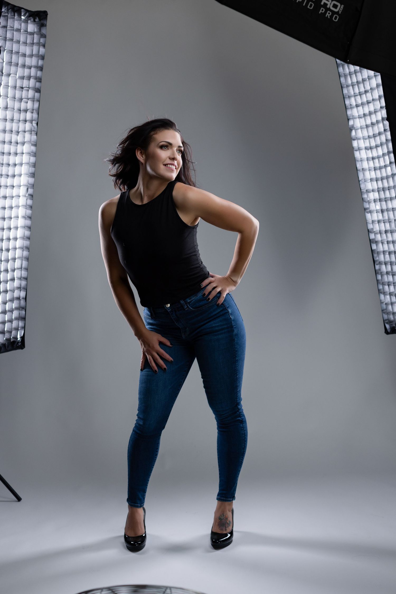 A model in black top and jeans poses dynamically in a professional photo studio with white backdrop and lighting setup.