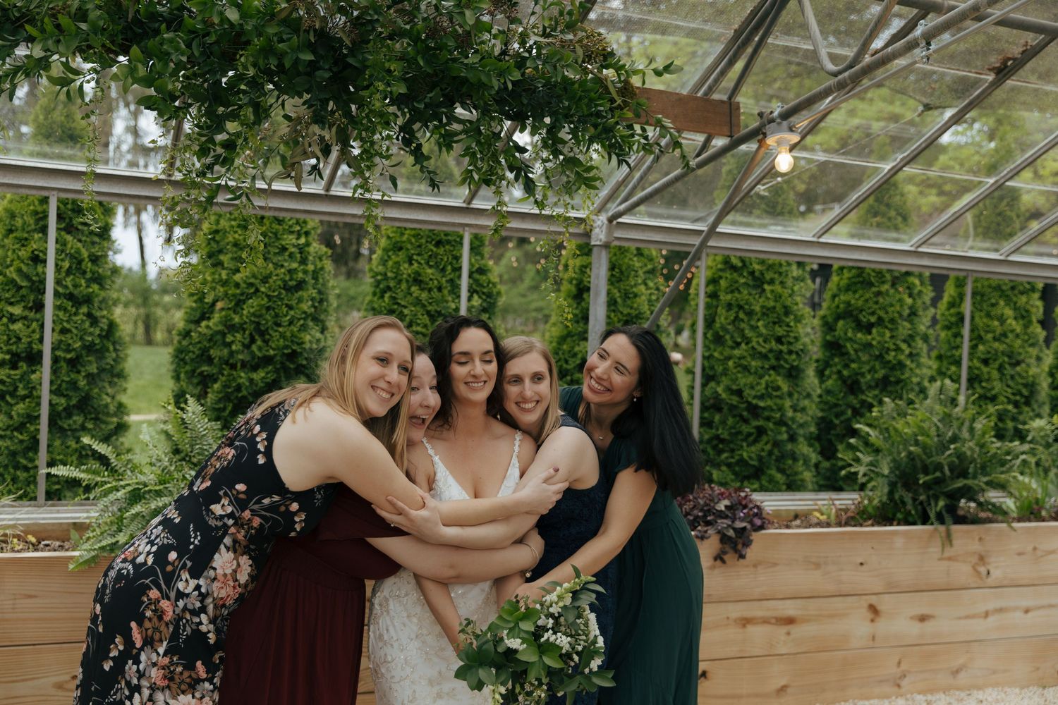 A group celebrates together in a sunlit greenhouse venue decorated with natural wood and greenery for a wedding event.