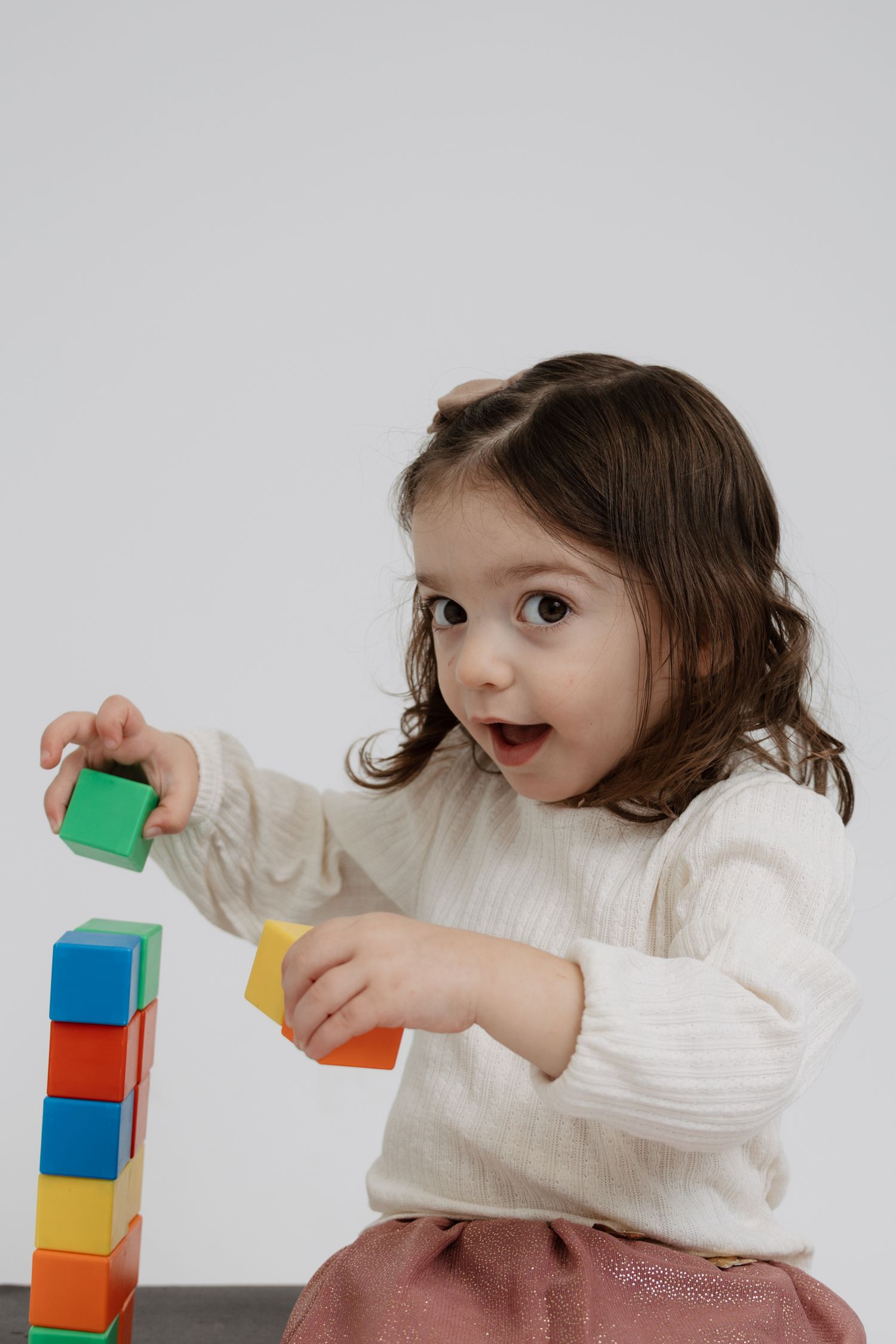 Child in white sweater plays with colorful wooden building blocks against light gray background.