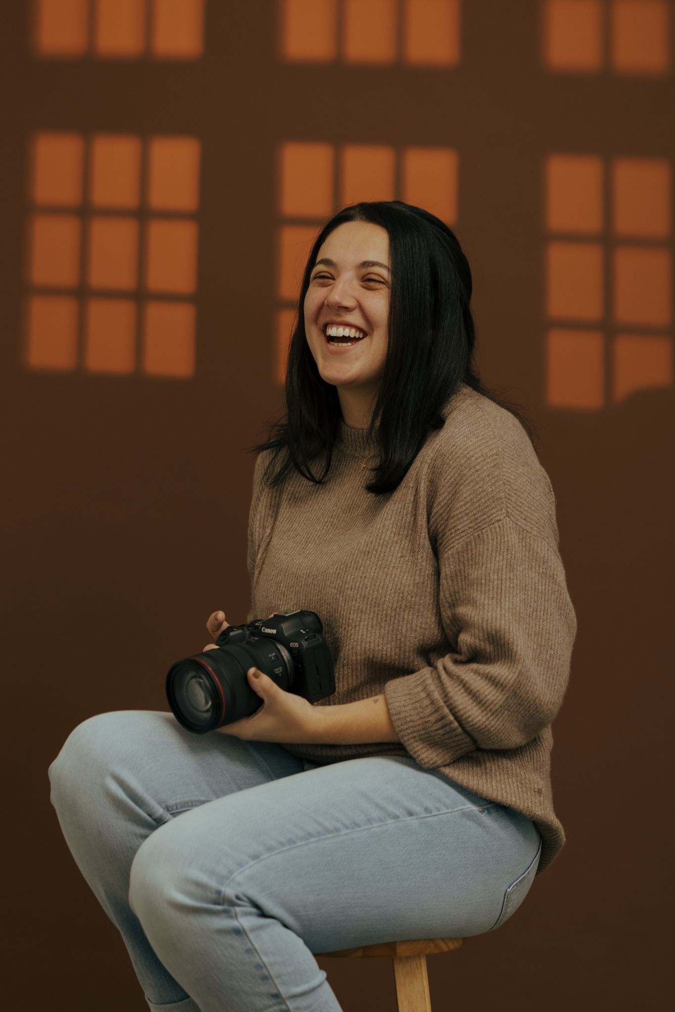 A person in a beige sweater and jeans sits casually with a camera against a warm orange-tinted background.