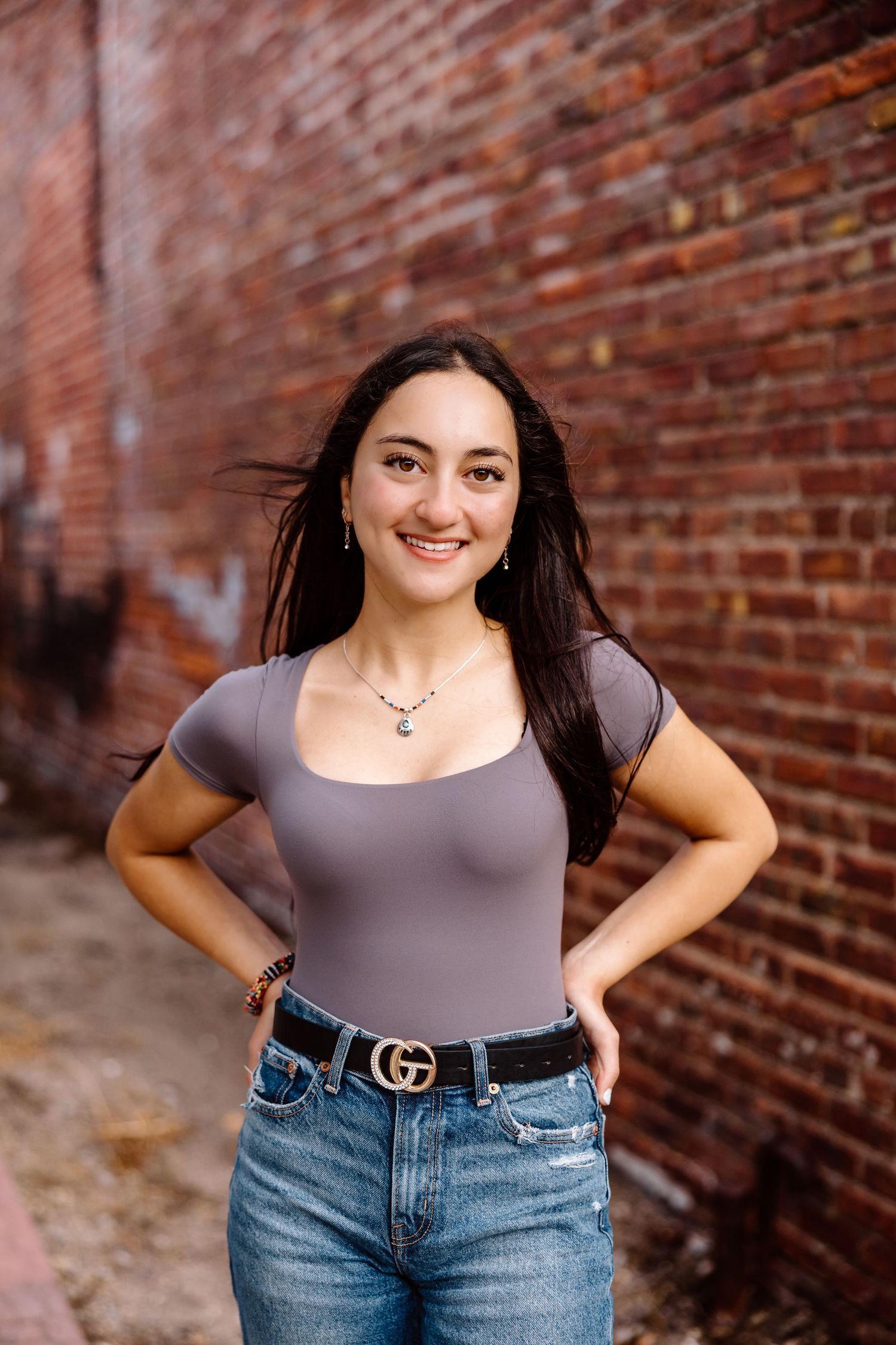 A person in a gray top and blue jeans poses against a rustic brick wall in an urban setting.